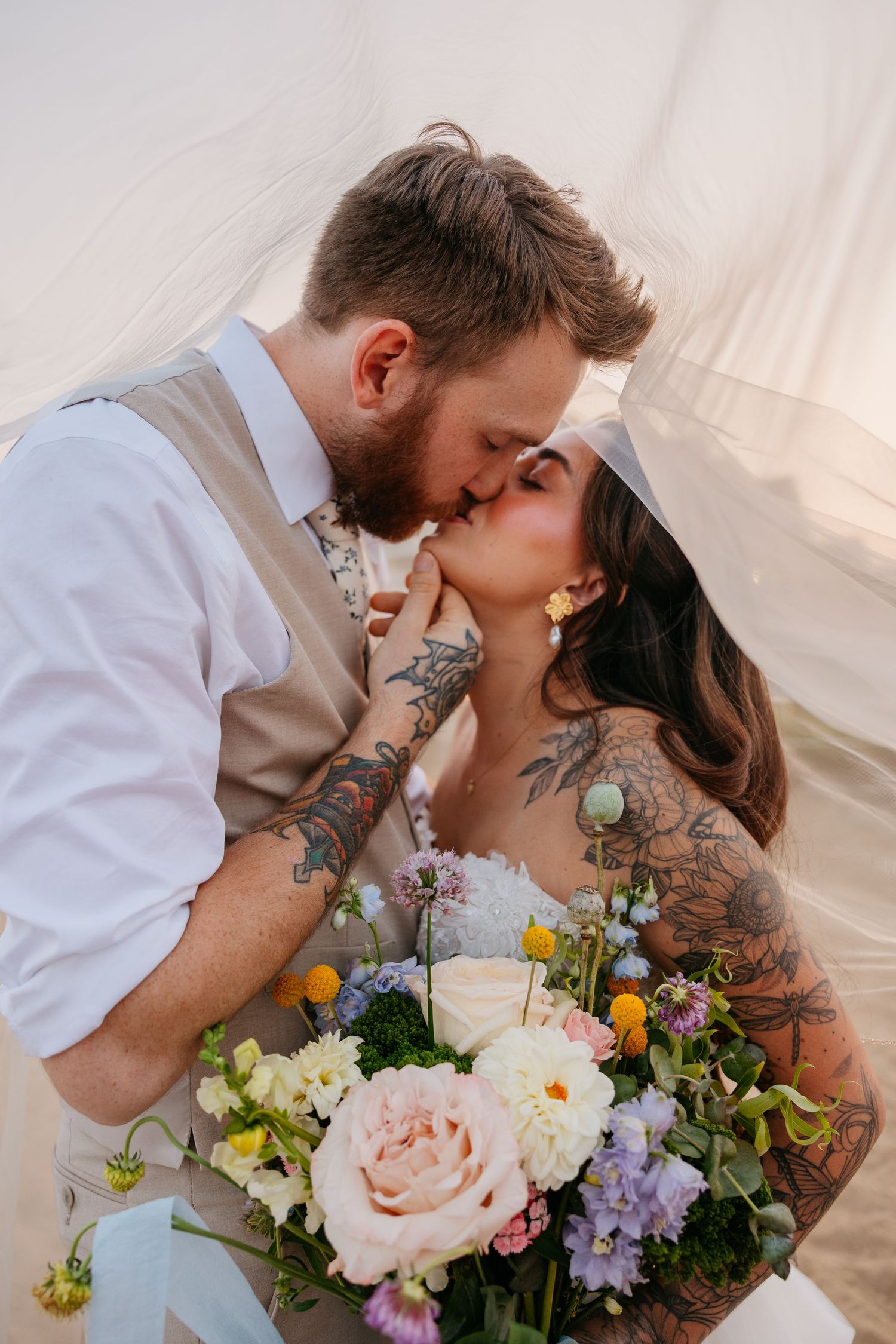 Tattooed bride and groom sharing a romantic kiss under a veil, bride holding a colorful wildflower bouquet.
