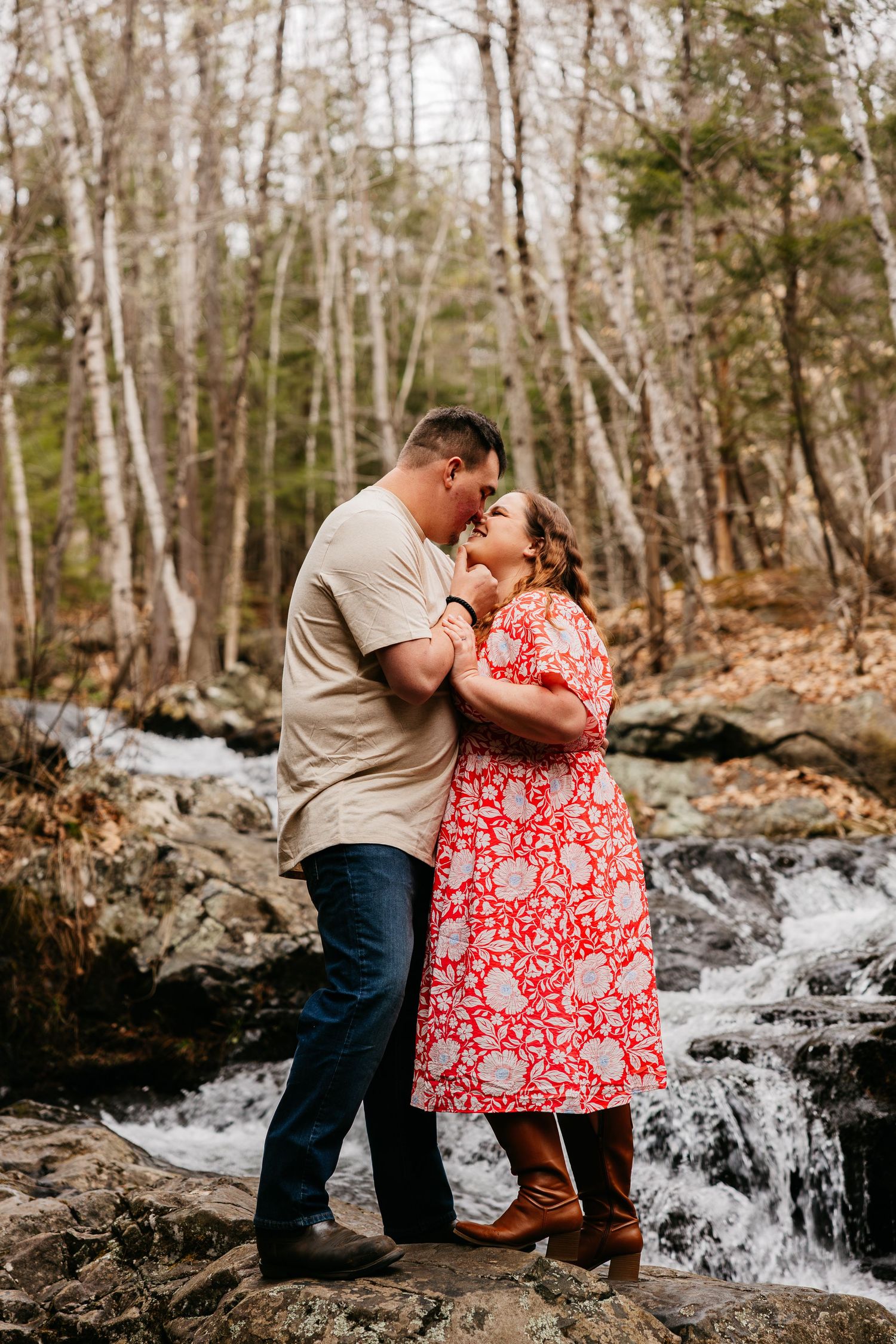 Couple sharing a romantic moment by a woodland waterfall, woman in red floral dress and man in beige shirt embracing on rocks.
