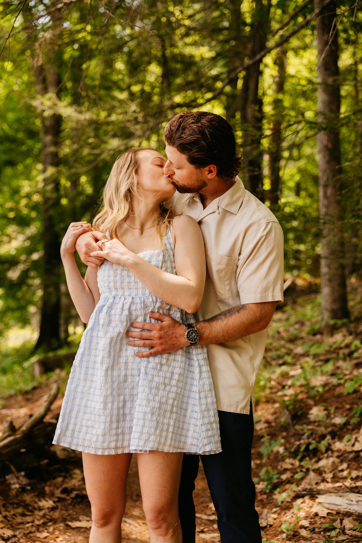 A couple shares a romantic kiss in a sunlit forest, the woman in a blue sundress and man in a beige shirt embracing tenderly.