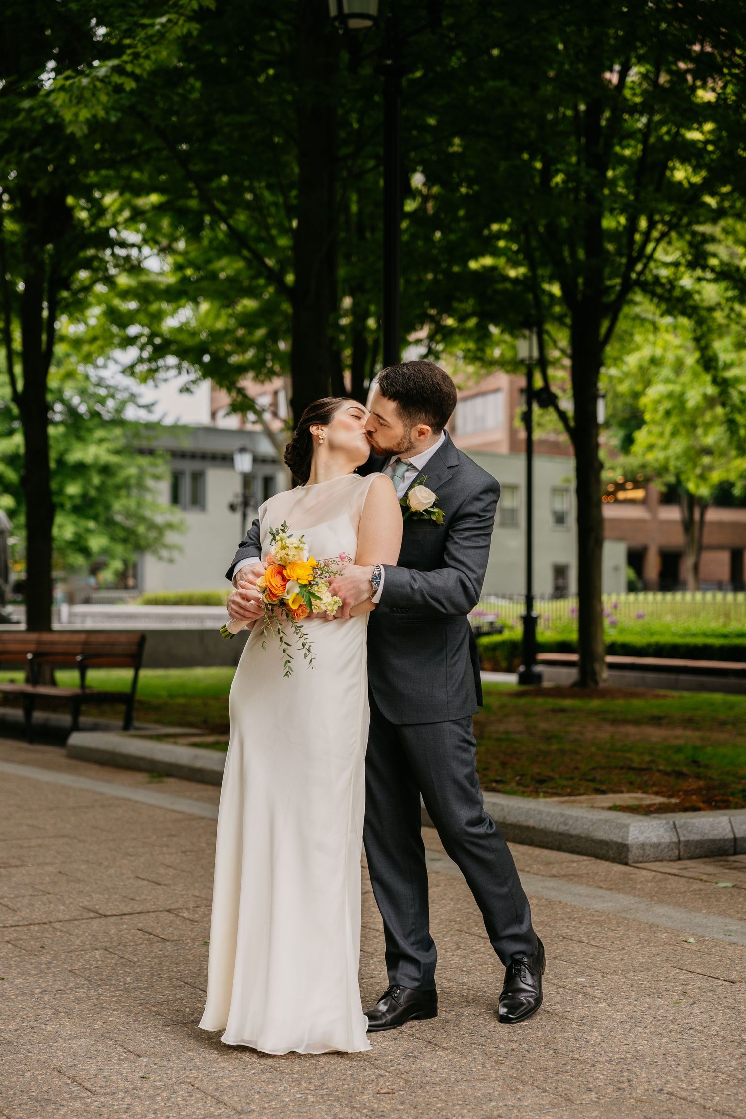 A bride and groom share a romantic kiss in a lush green park, she holds a vibrant orange and yellow bouquet.