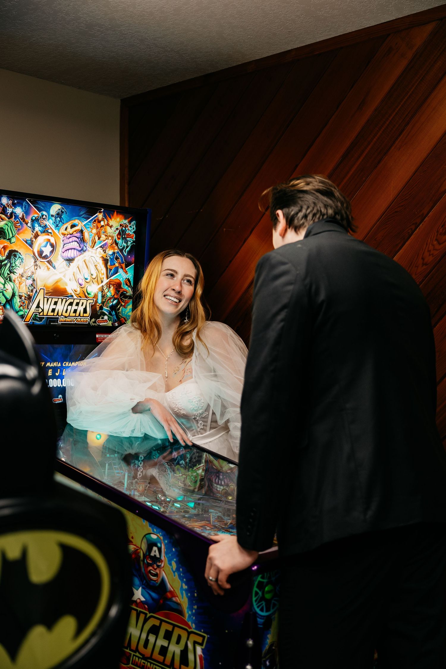 A couple plays arcade games together in a dimly lit room with colorful game screens visible in the background.
