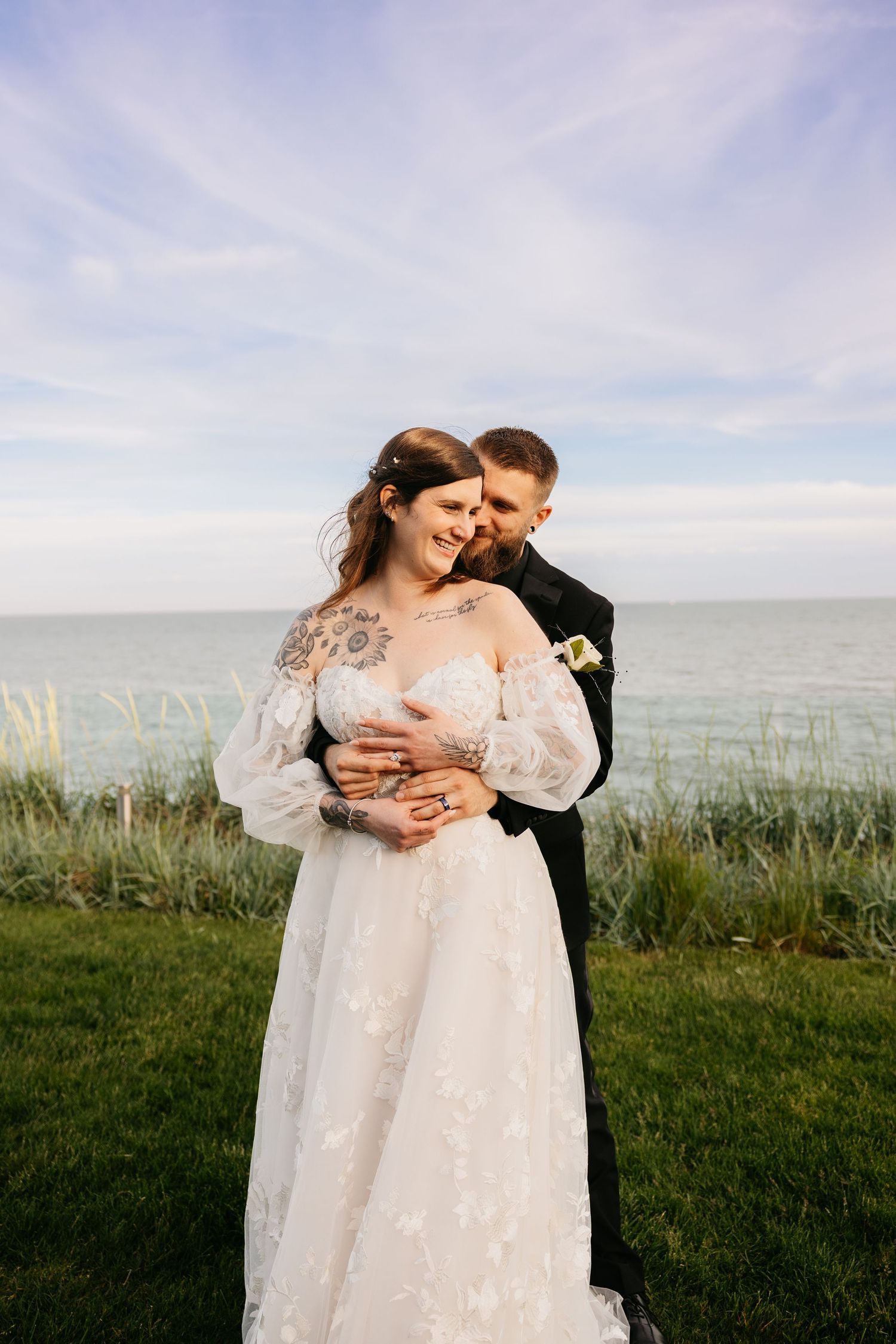 Groom embraces bride in floral lace wedding gown with tattoos, standing on green cliff overlooking ocean under blue sky.