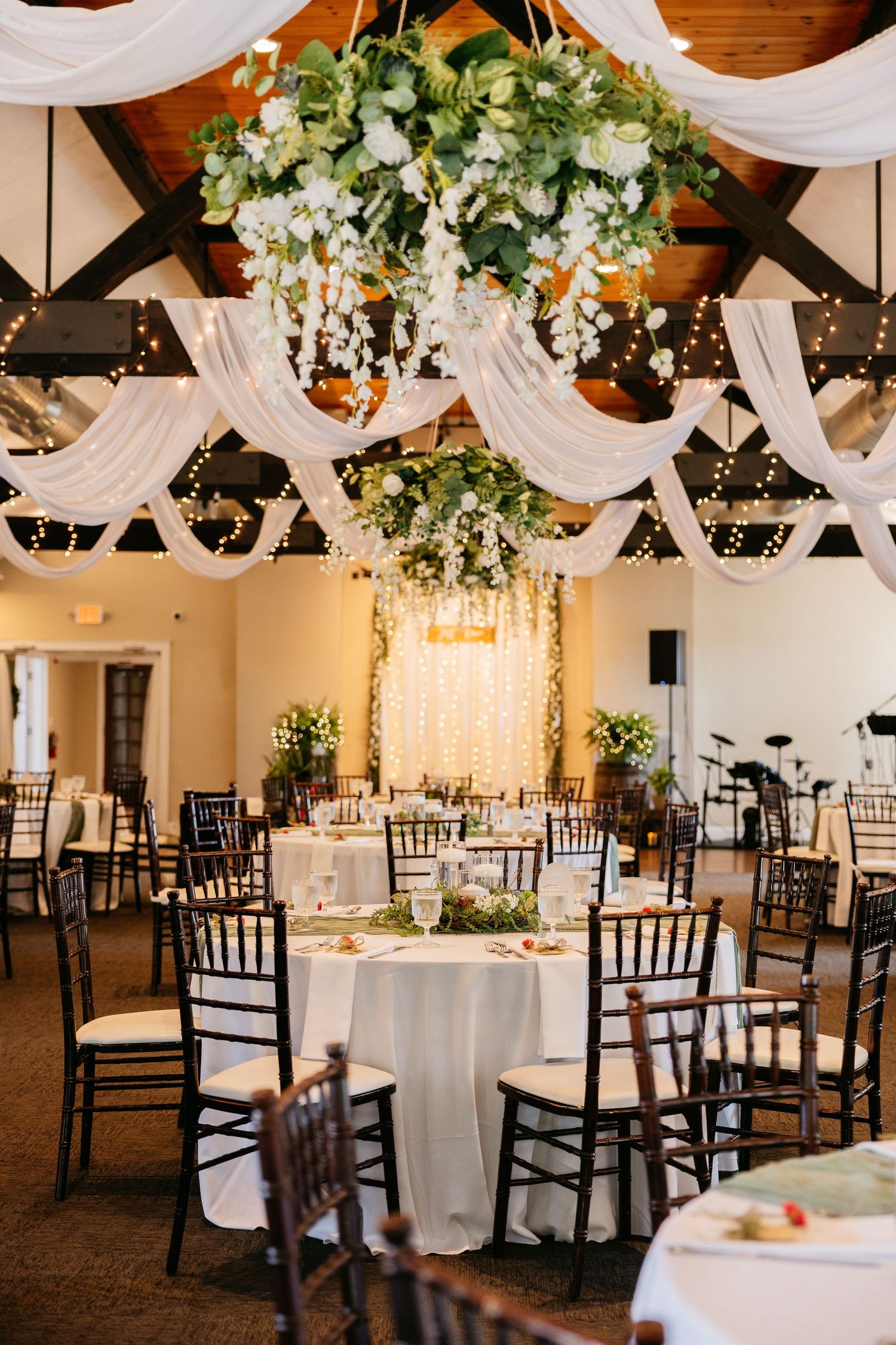 Elegant wedding reception hall with floral chandelier, white draping, string lights, and dark Chiavari chairs around round tables.