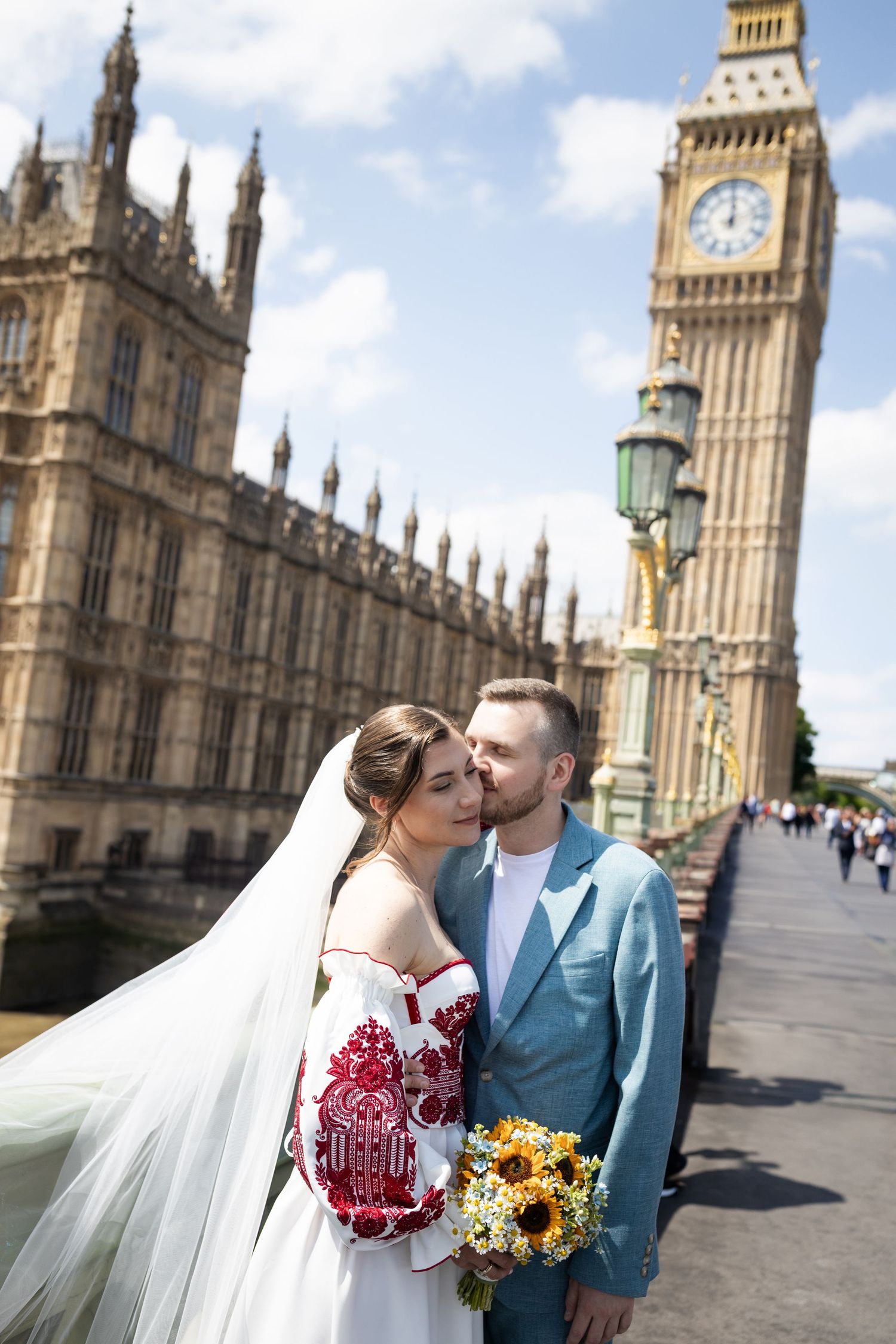 Bride and groom pose on Westminster Bridge with Big Ben behind them, bride wearing red embroidered gown holding sunflowers.