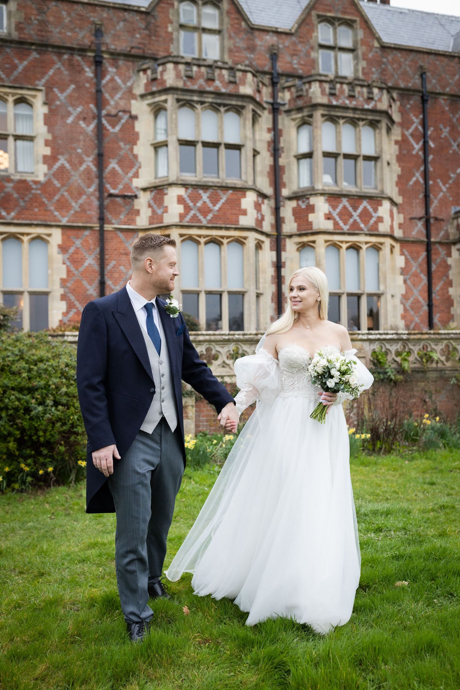Bride and groom holding hands walking in front of a grand red brick manor house on their wedding day.