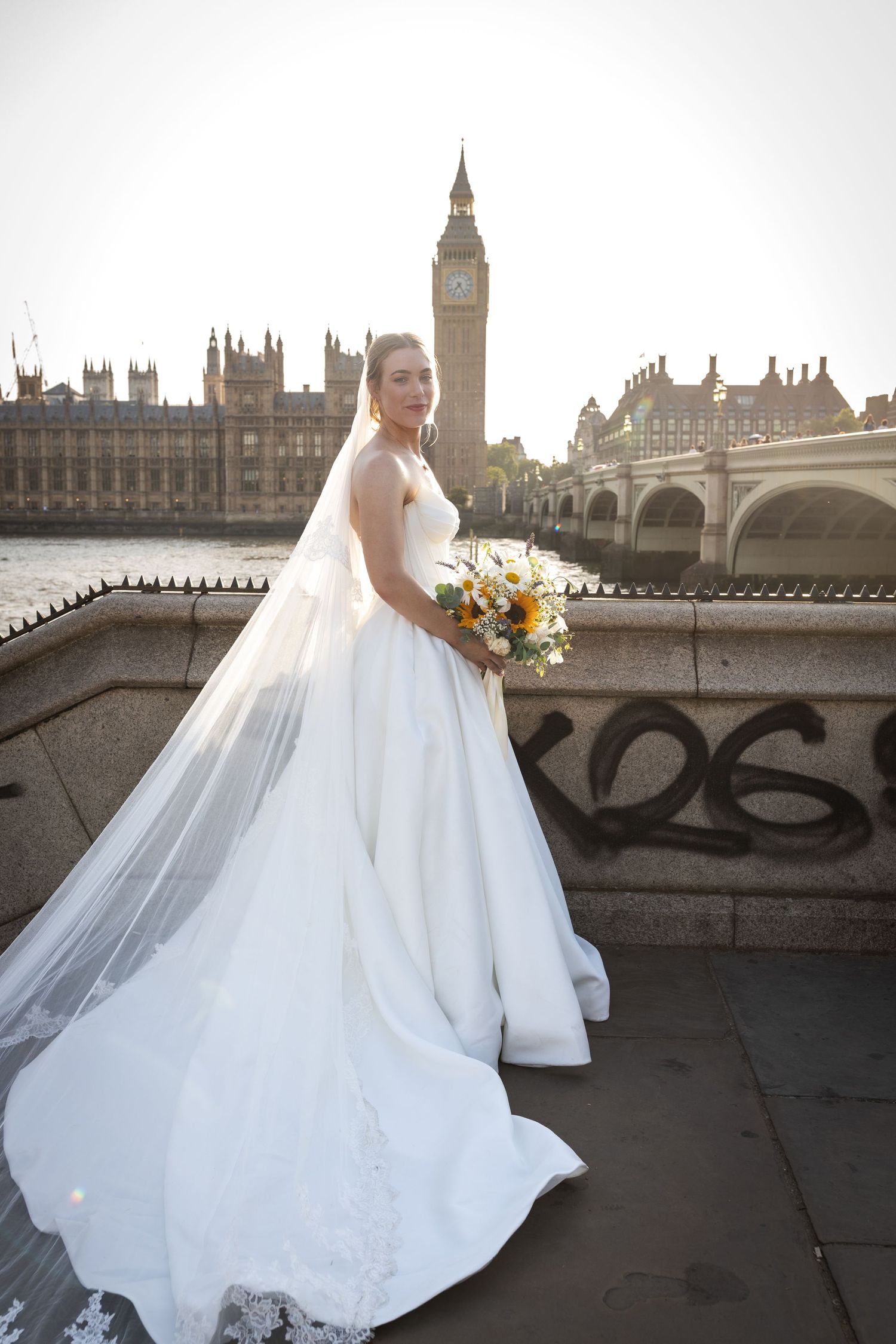 Bride in white gown with veil holds bouquet by Westminster Bridge with Big Ben and Houses of Parliament, London.