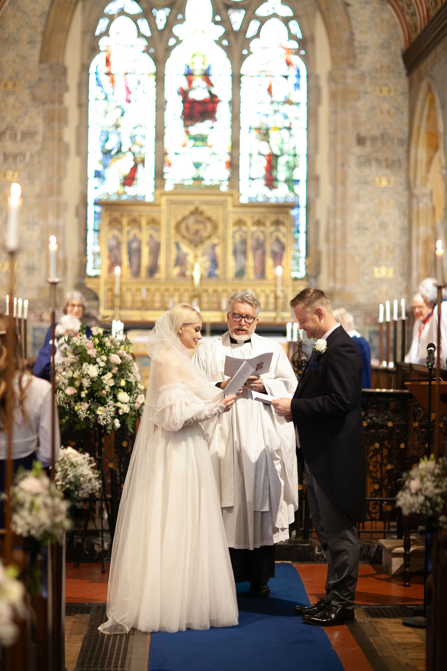 Bride and groom exchange vows at church altar with officiant, stained glass windows and floral arrangements in background.