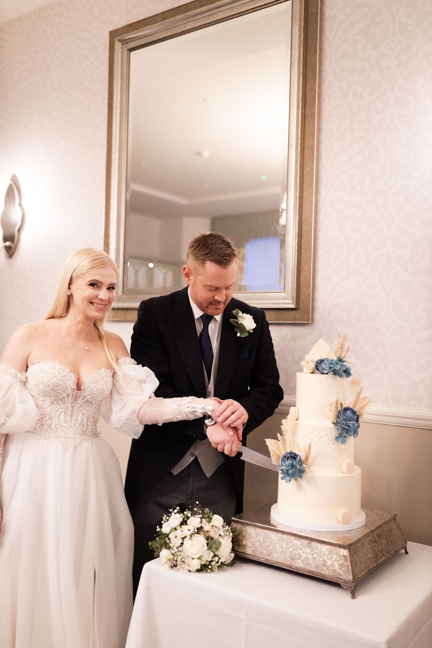 Bride and groom cutting a three-tier white wedding cake with blue floral decorations at their elegant wedding reception.