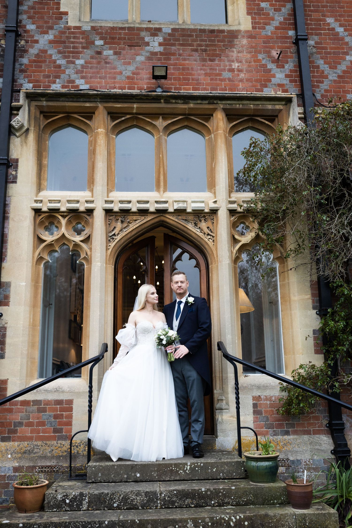Bride and groom pose on stone steps outside a Gothic-style red brick manor house on their wedding day.