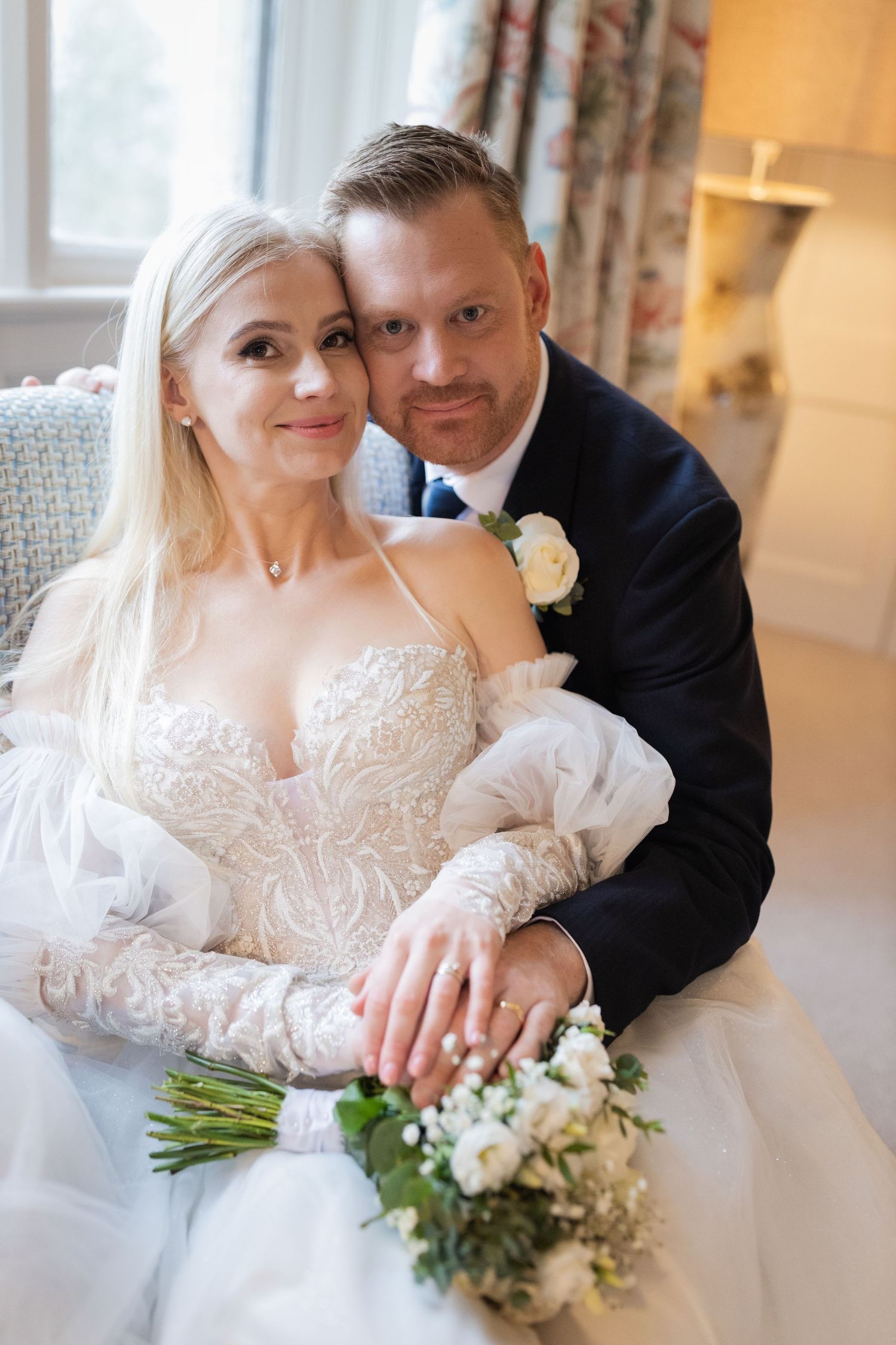Bride and groom pose together on a chair, bride in lace gown holding white bouquet, groom in navy suit with rose boutonniere.