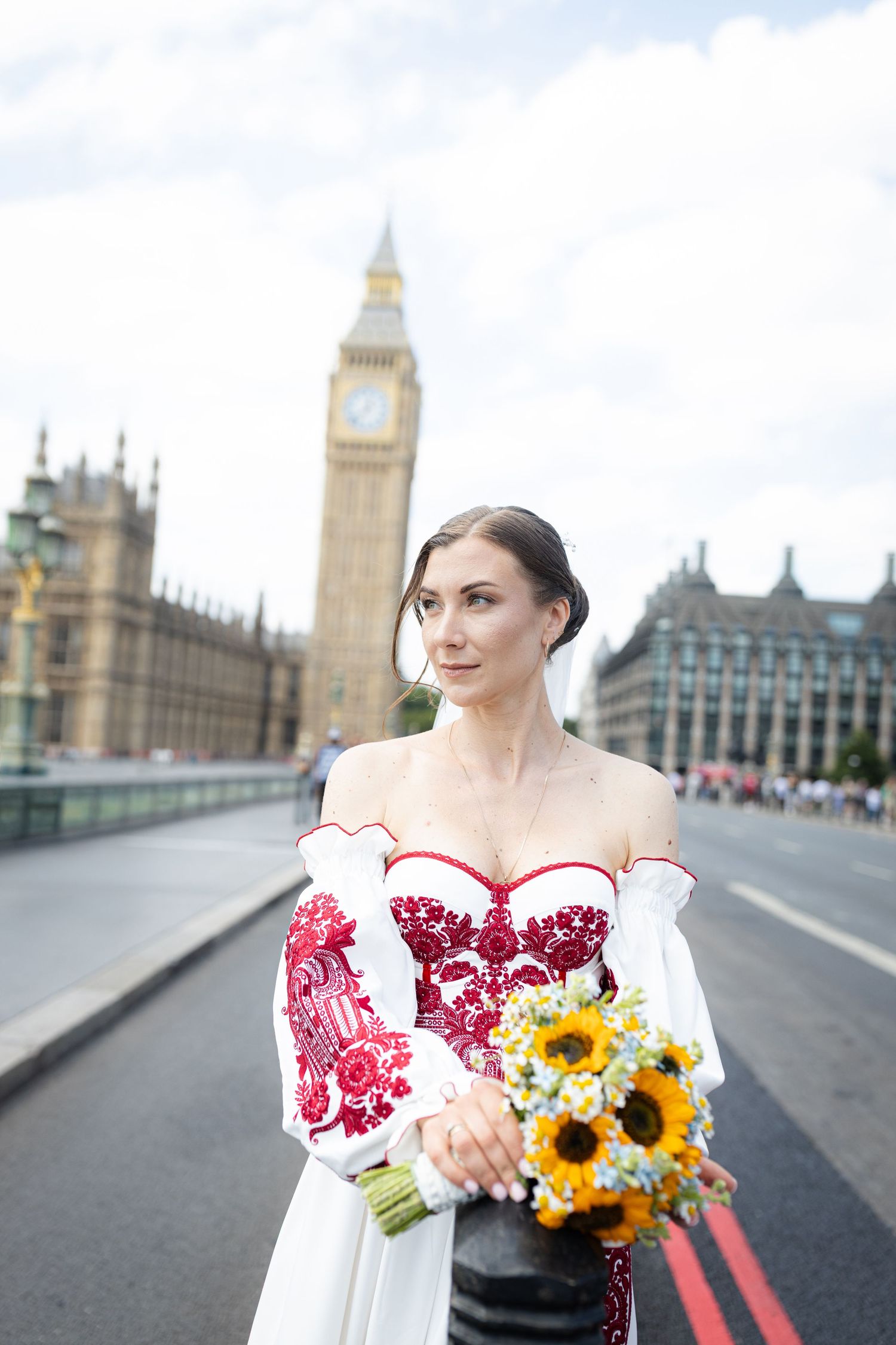 Bride in red embroidered white gown holds sunflower bouquet on Westminster Bridge with Big Ben backdrop in London.