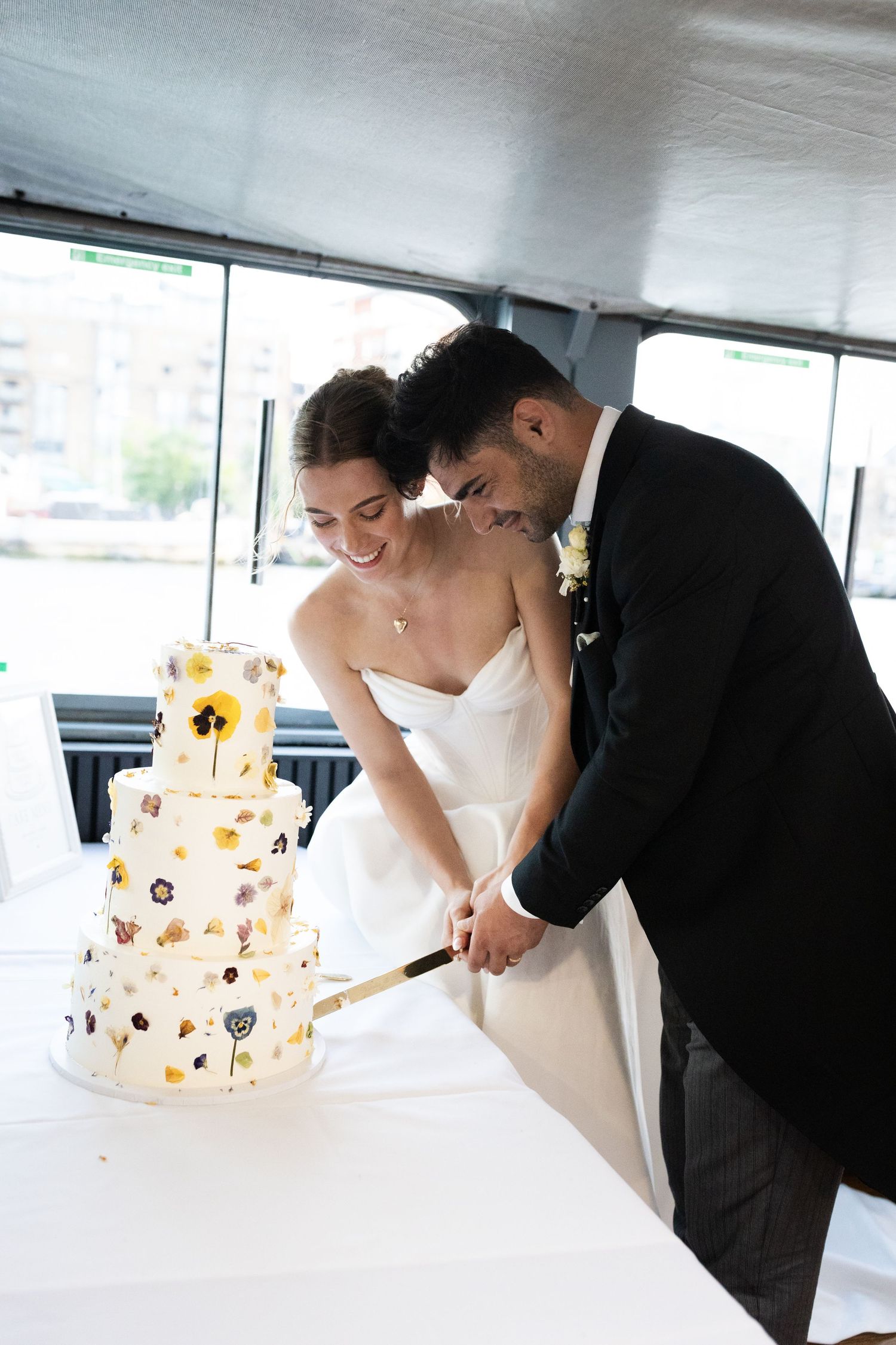 Bride and groom cutting their white floral wedding cake together on a boat venue, smiling joyfully.
