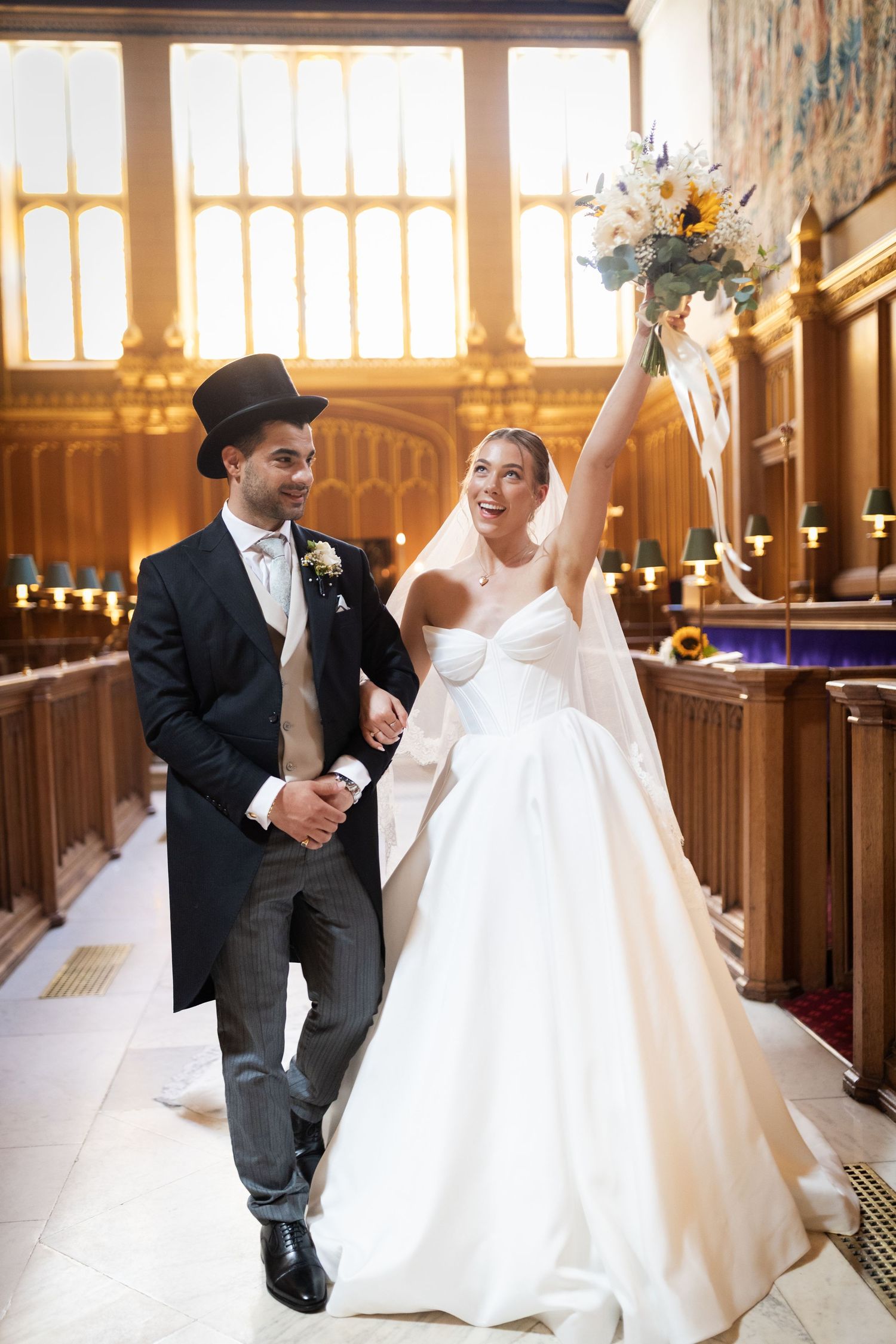 Joyful newlyweds walk down church aisle, bride in white gown raising sunflower bouquet, groom in black top hat and tails.