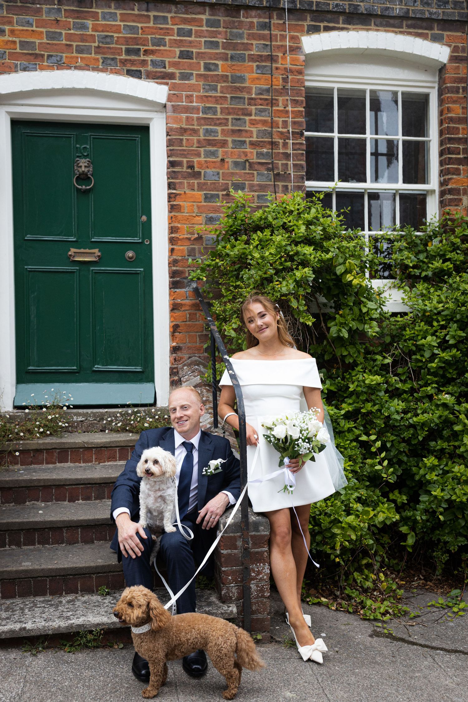 A couple poses outside a brick house with their small dogs in a series of casual photos by a green front door.