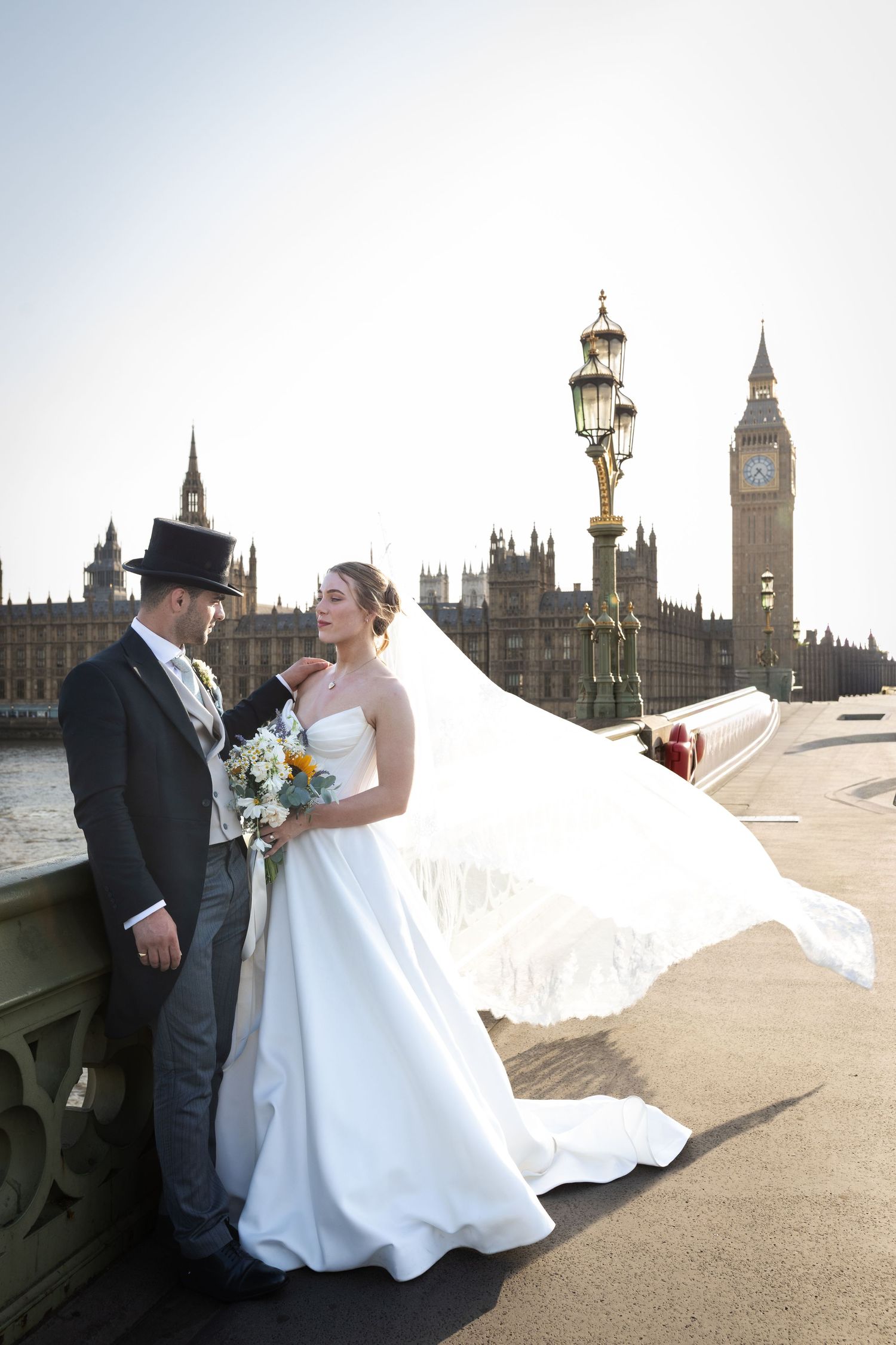 Bride and groom pose on Westminster Bridge with Big Ben and Houses of Parliament backdrop, veil flowing in wind.