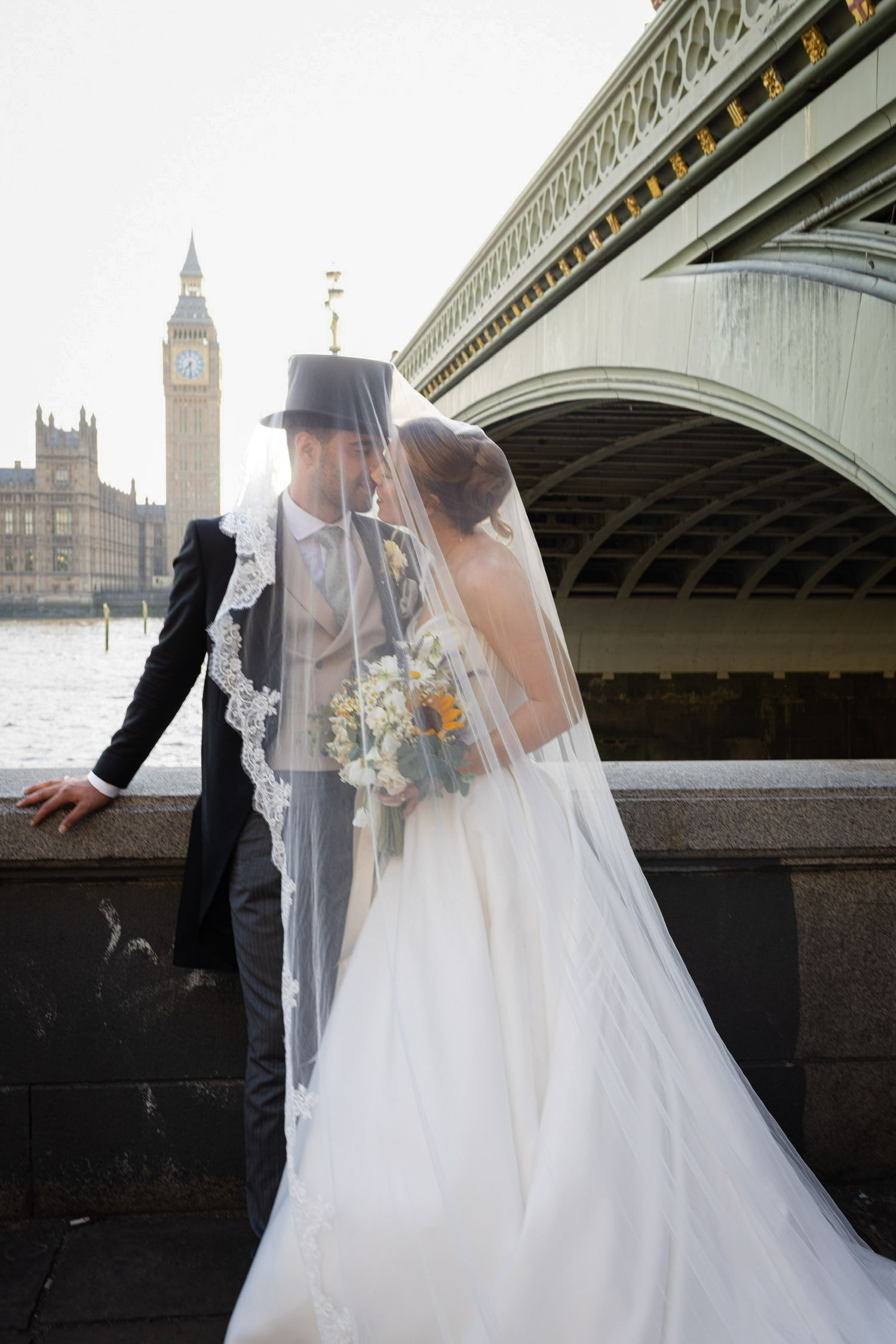 Newlyweds sharing a kiss under bridal veil near Westminster Bridge with Big Ben in background.