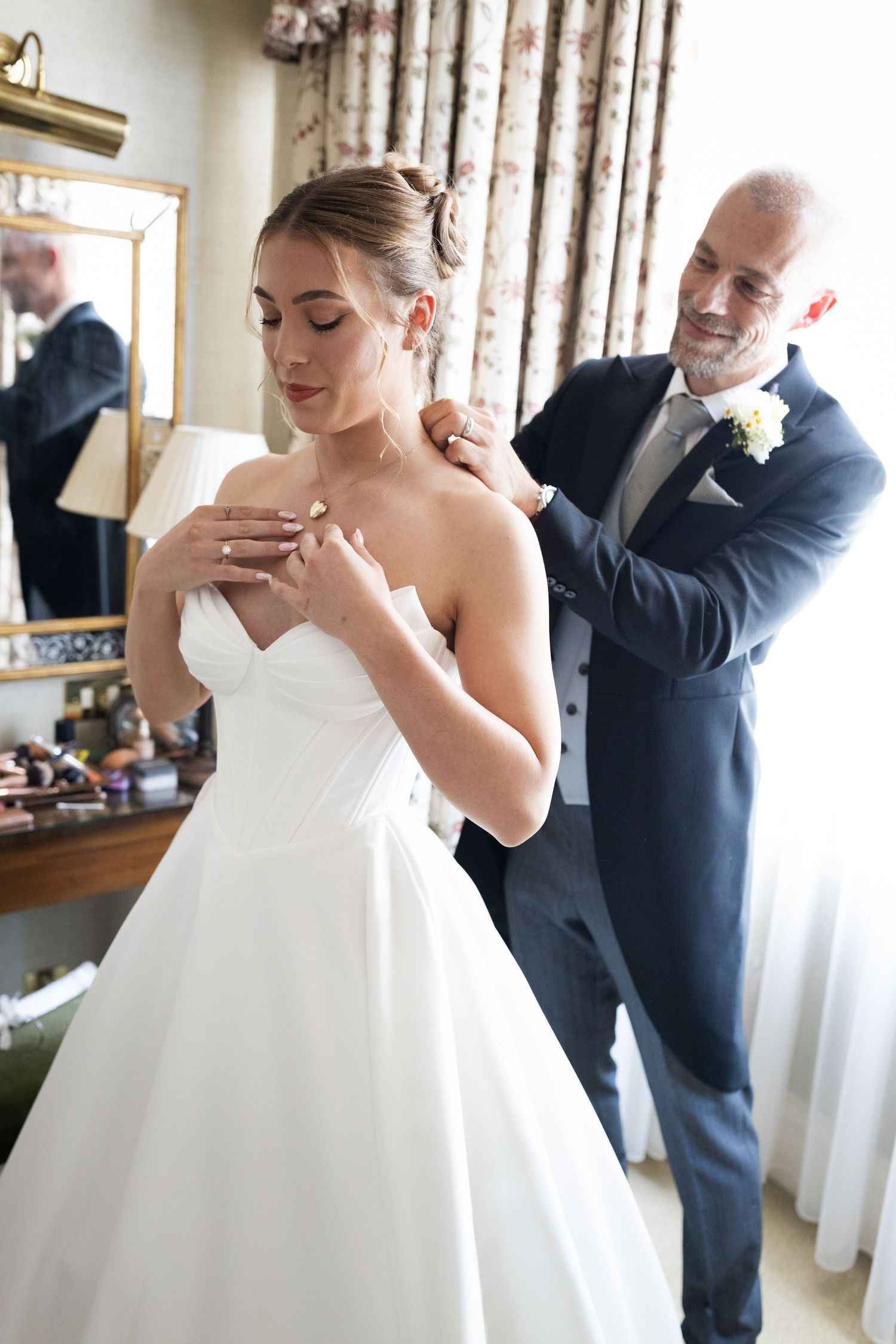 Father of the bride helping daughter with necklace in bridal suite before wedding ceremony.