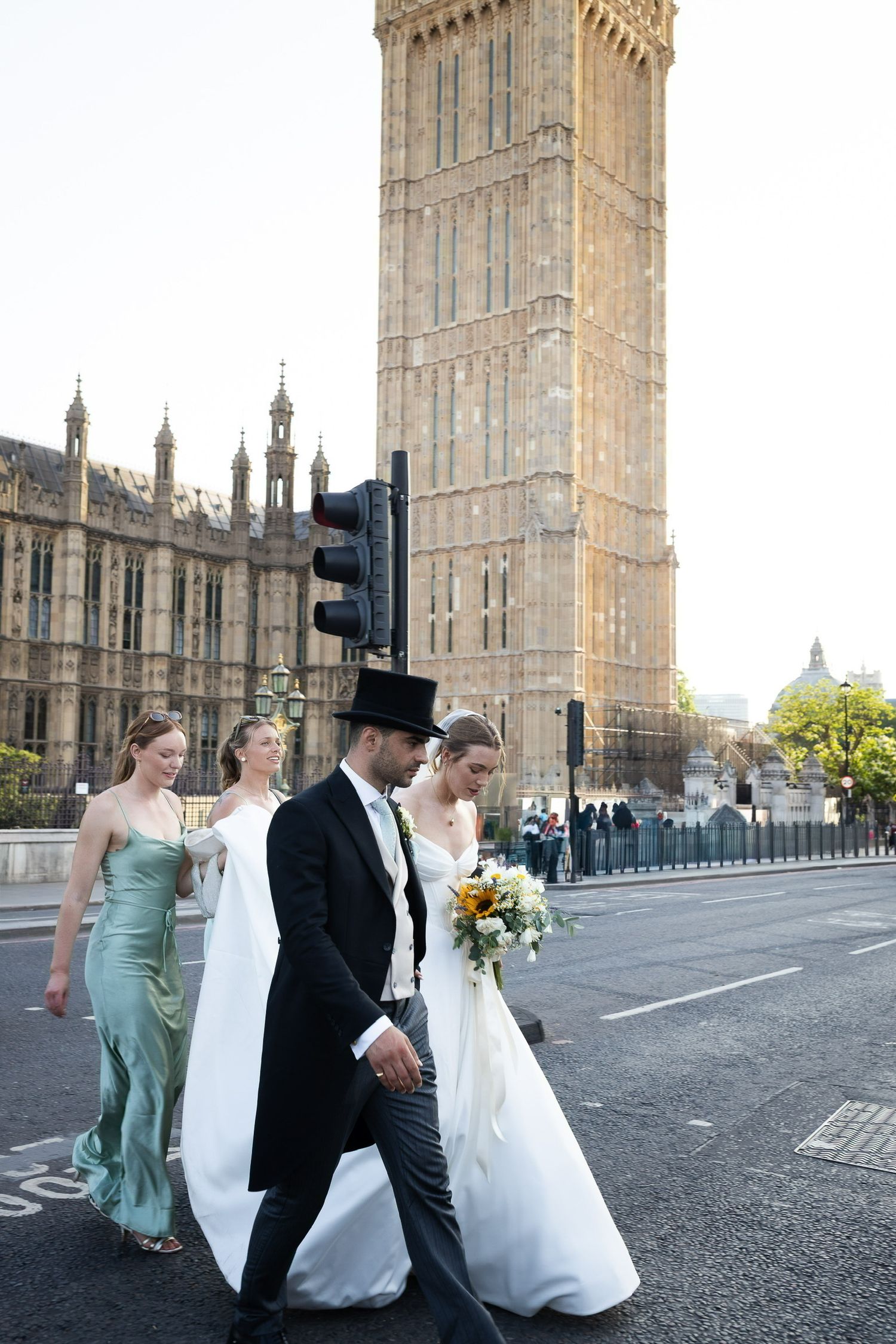 Wedding couple walks past Big Ben in London, bride in white gown with bouquet, groom in top hat and tails.