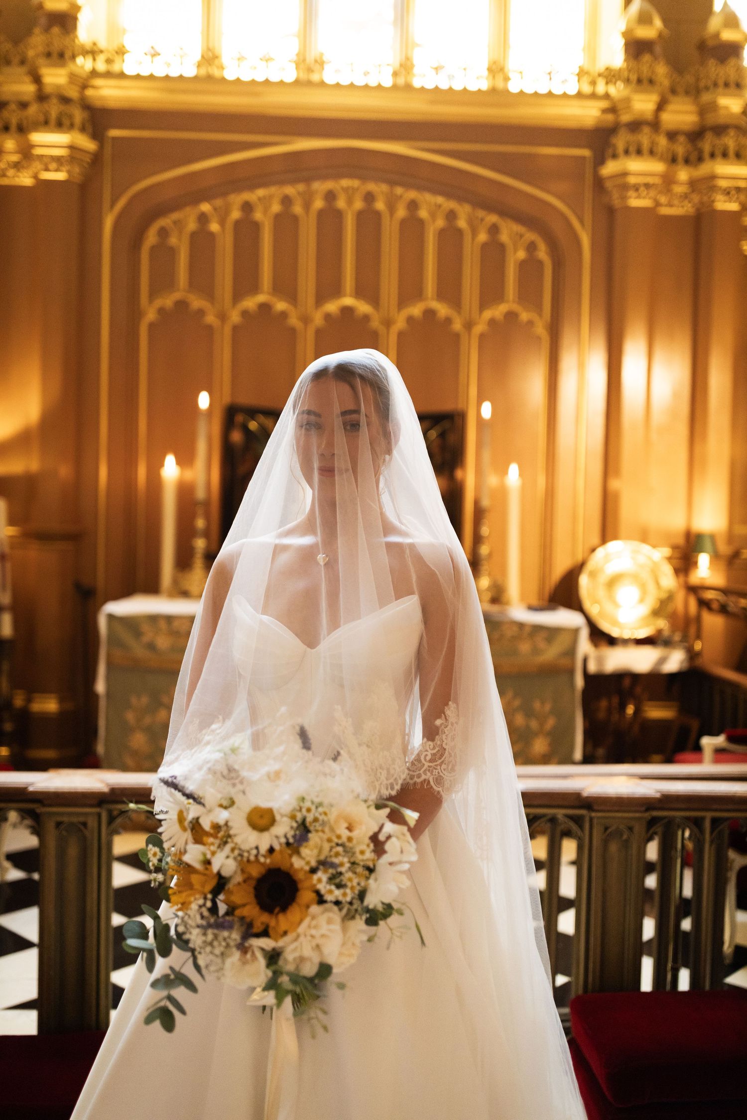 Bride in white gown and veil holds sunflower bouquet inside ornate golden chapel with candles.