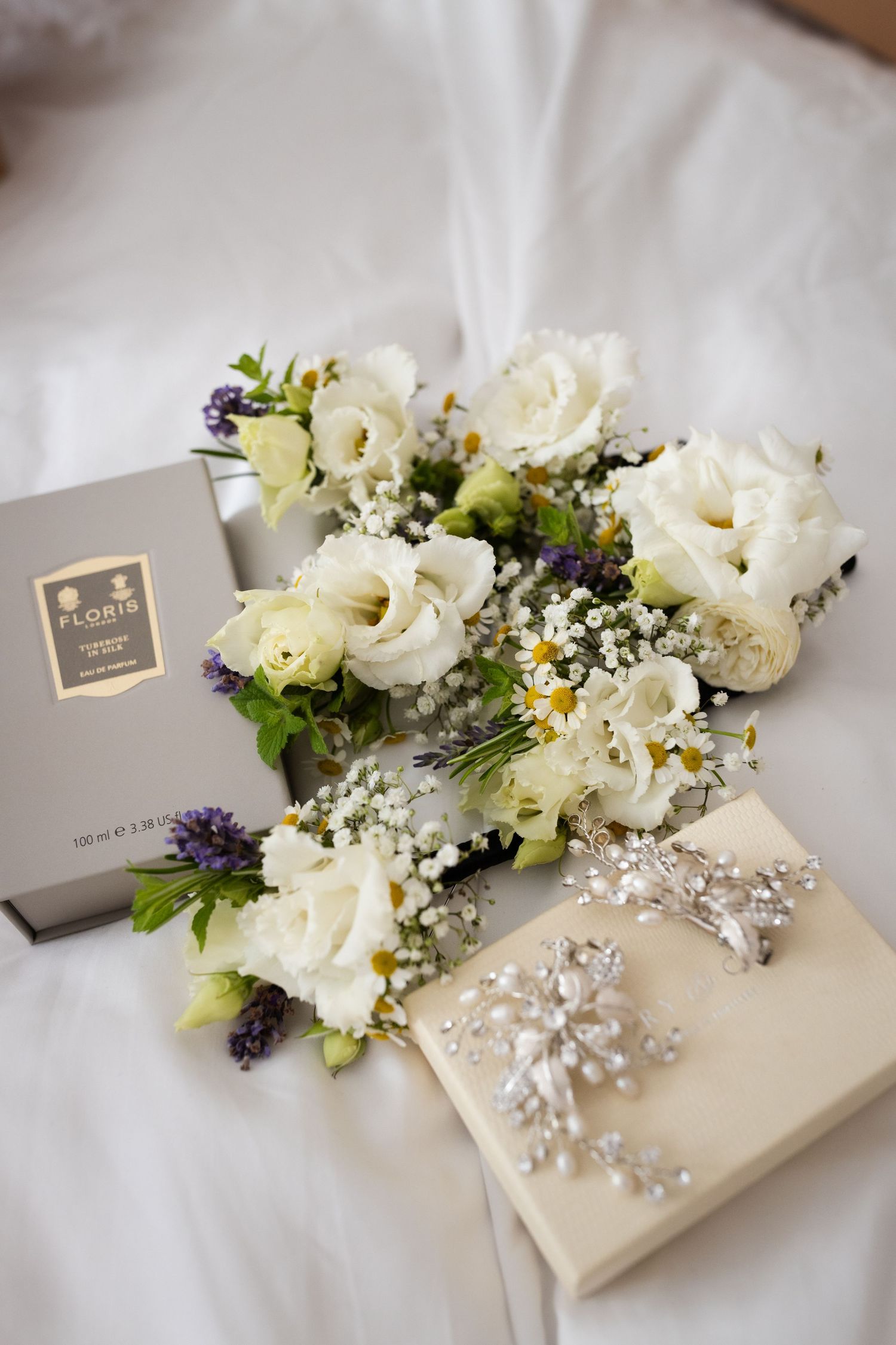 Elegant wedding flat lay with white roses, lavender, gypsophila, Floris perfume box, and crystal hair accessory on white fabric.