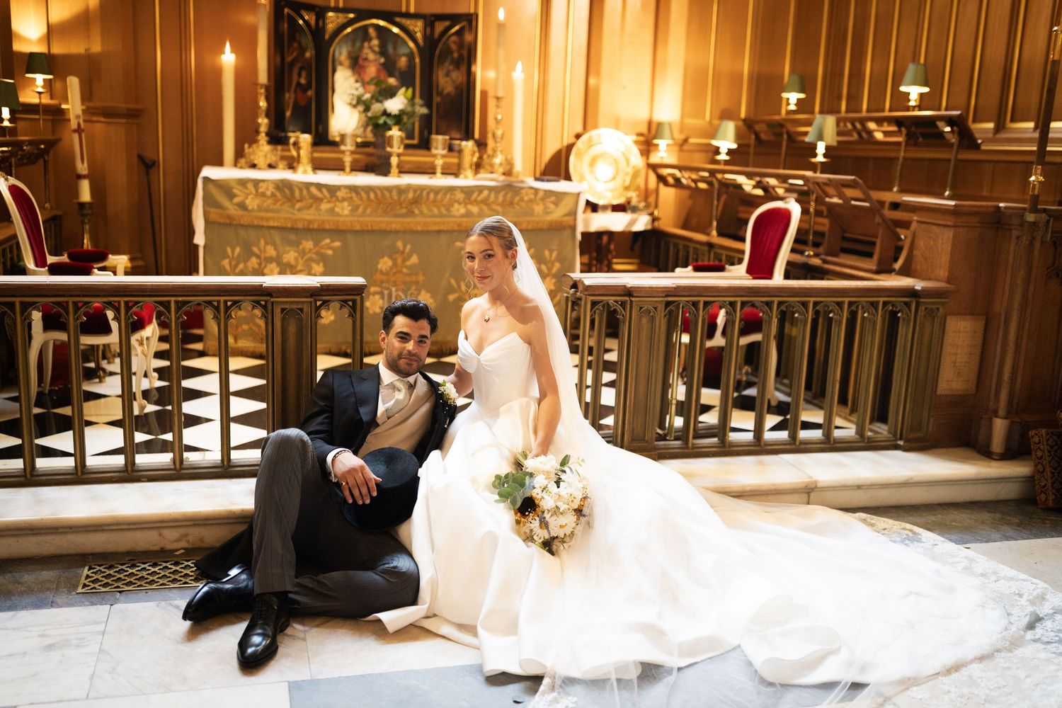 Bride and groom posing together on church altar steps, bride in ballgown holding bouquet, groom in dark suit.