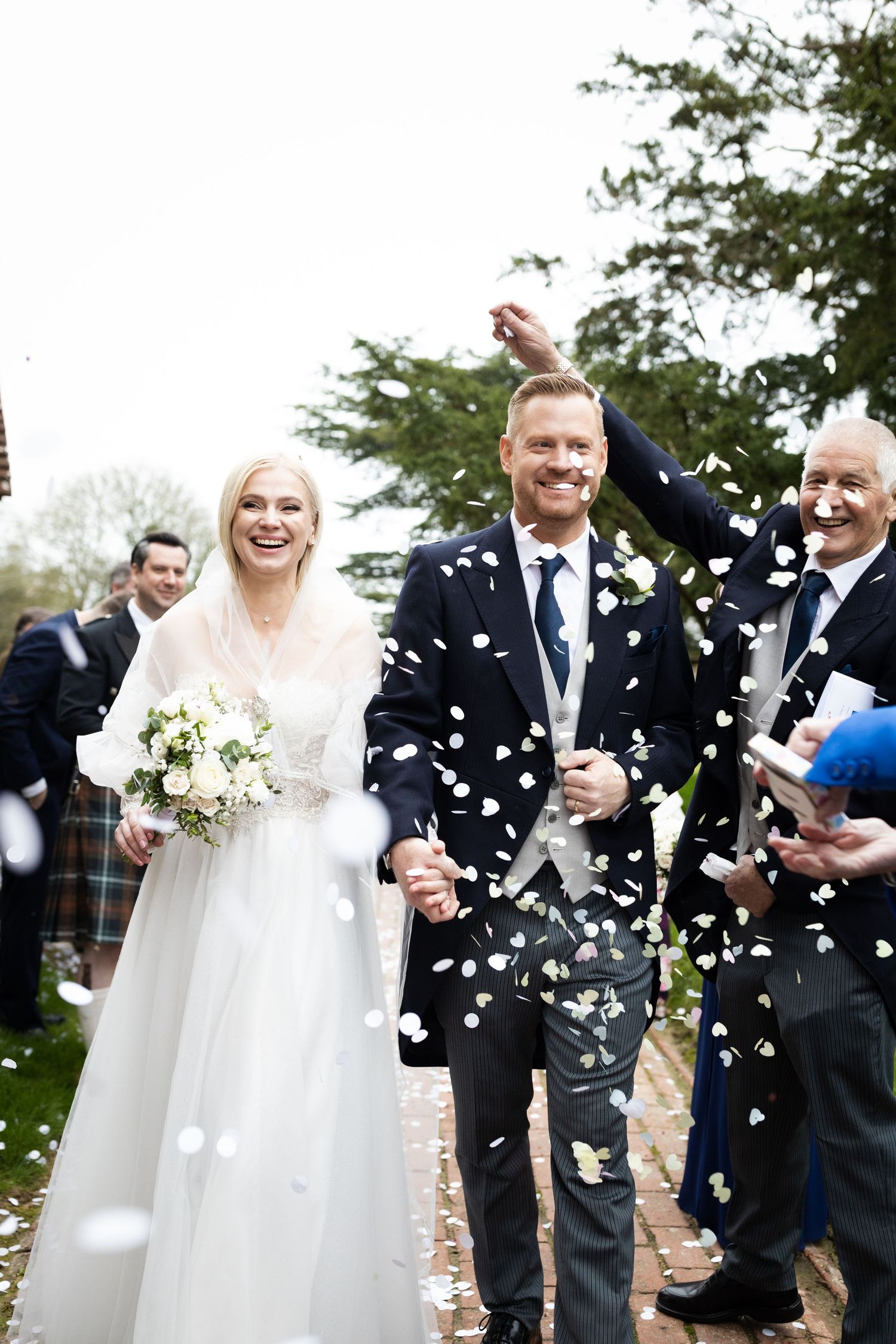 Joyful newlyweds walk through confetti shower, bride in white gown holding bouquet, groom in dark suit, guests celebrating outdoors.