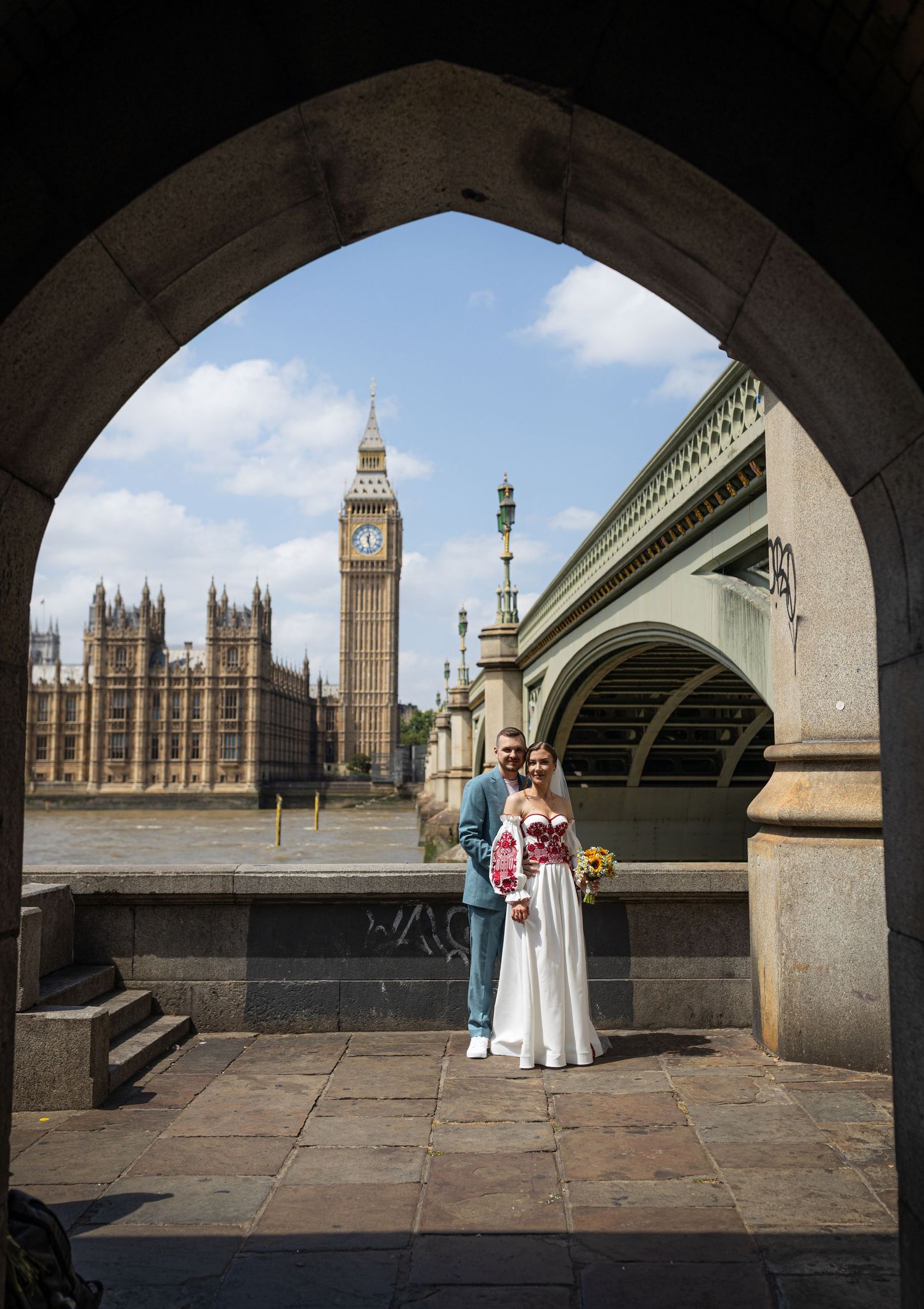 Couple poses under stone arch with Big Ben and Westminster Bridge in London backdrop on sunny day.