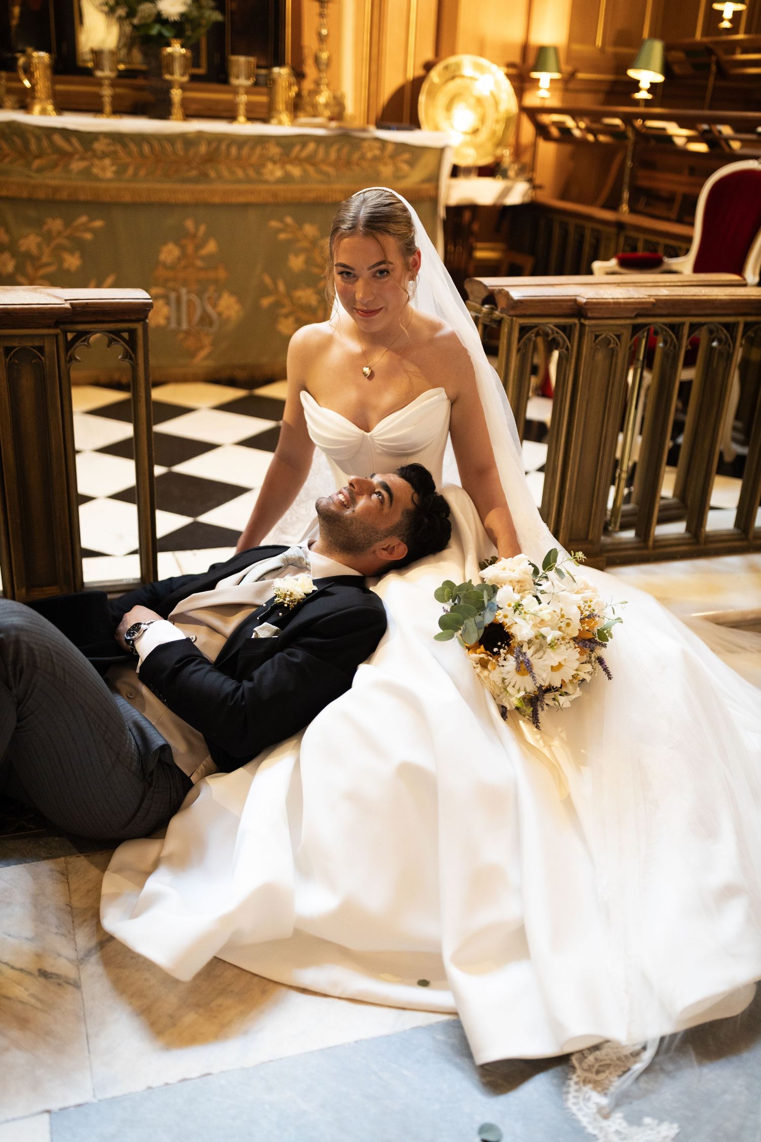 Bride in white gown sits on church floor with groom lying across her lap, holding a bouquet in an ornate venue.