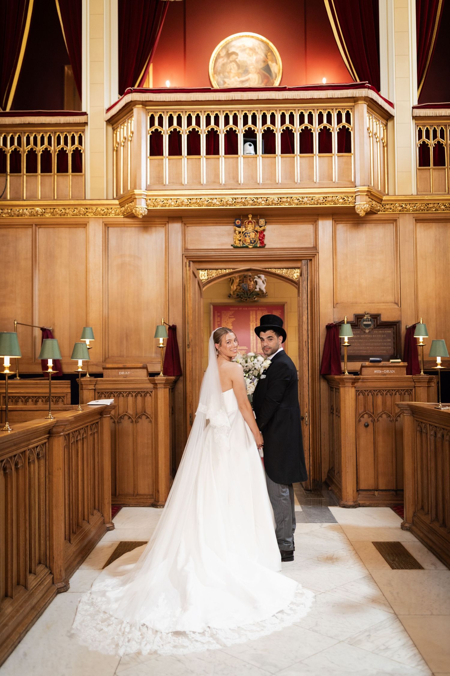 Bride and groom pose in elegant wood-paneled wedding venue with ornate balcony and gold details.
