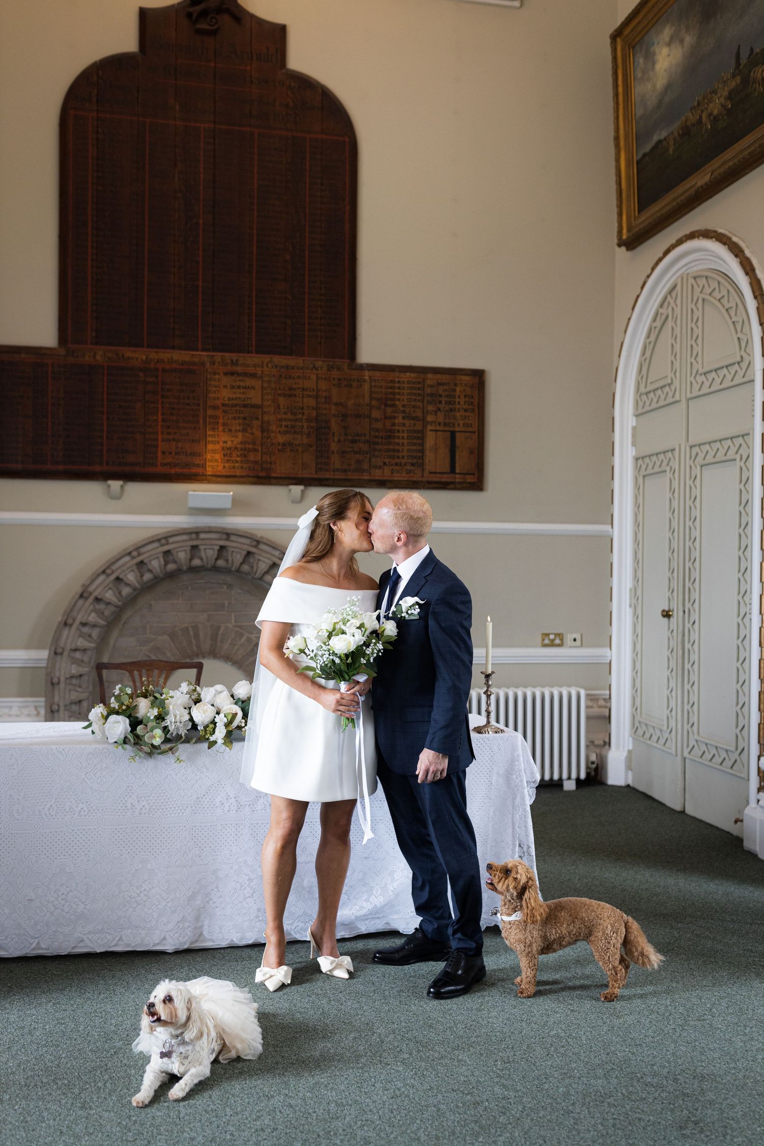 A couple shares a tender moment at their wedding ceremony with two dogs sitting beside them on green carpet.
