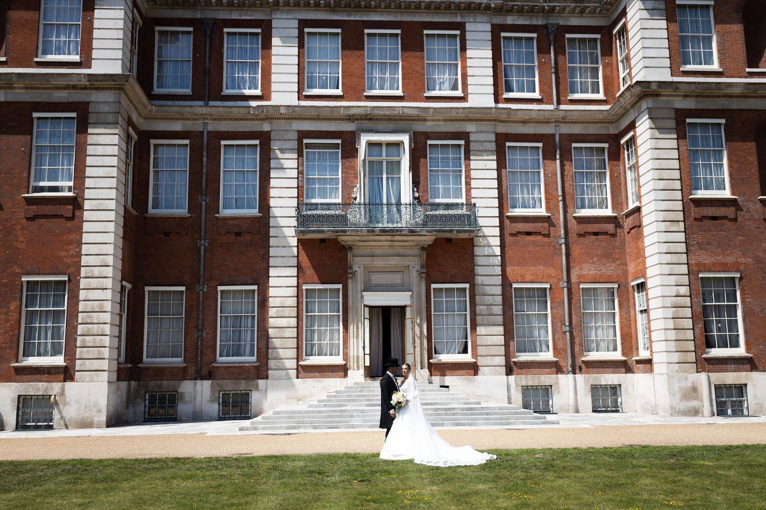 Bride and groom pose before a grand red brick Georgian manor house with white stone columns on a sunny day.