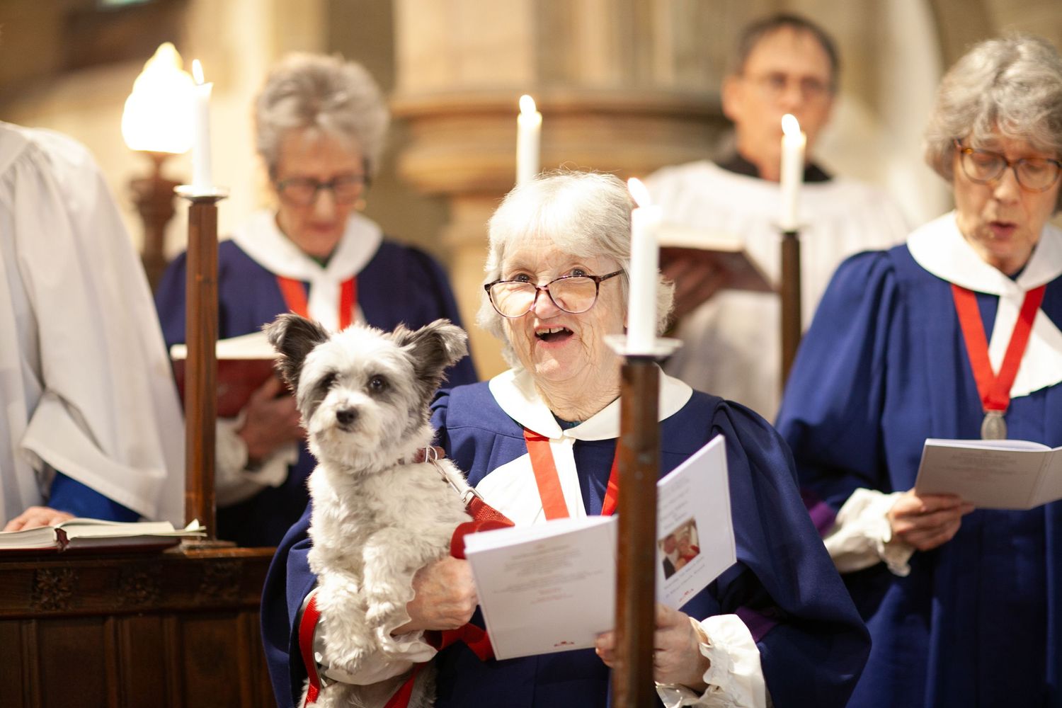 Church choir members in blue robes singing, with one woman holding a small gray and white dog during a service.