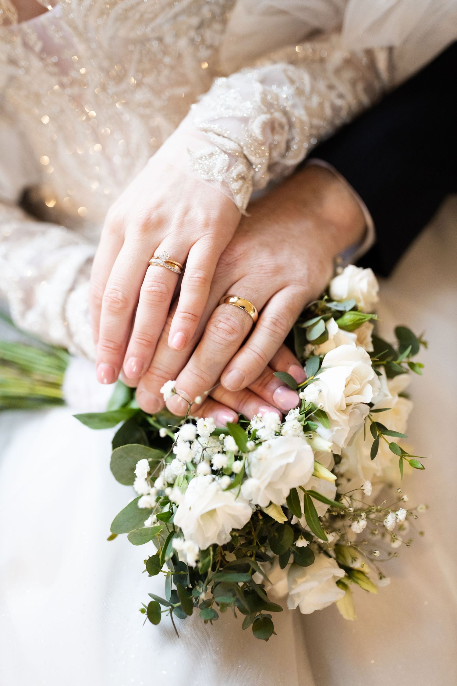 Newlywed couple displaying gold wedding rings on hands resting over a white bridal bouquet with roses and greenery.