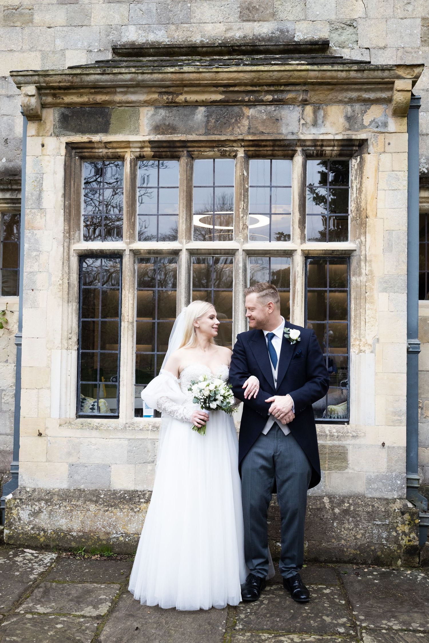 Bride and groom posing together outside a historic stone building with large windows on their wedding day.