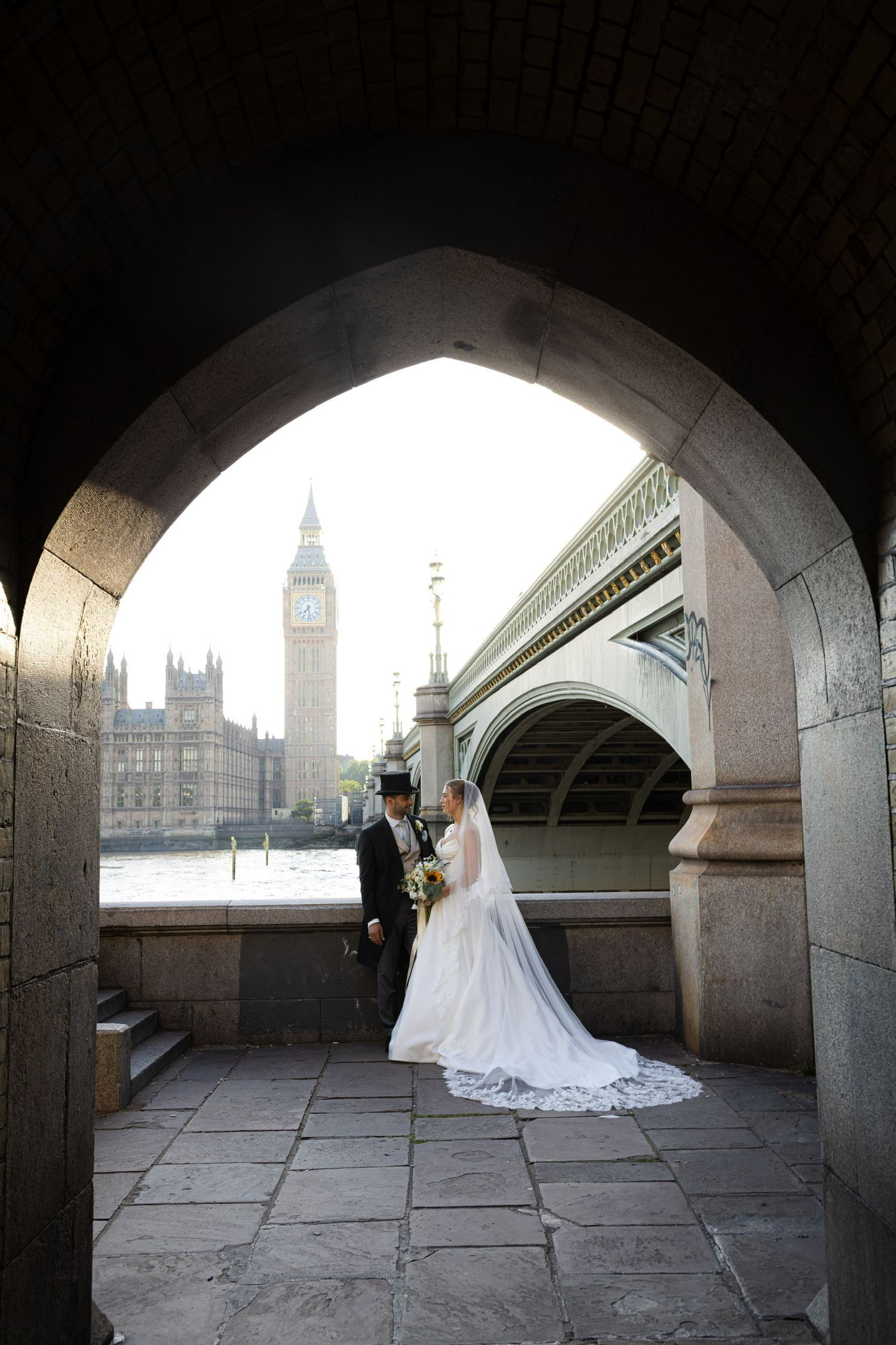 Bride and groom pose under stone arch with Big Ben and Westminster Bridge in London backdrop.