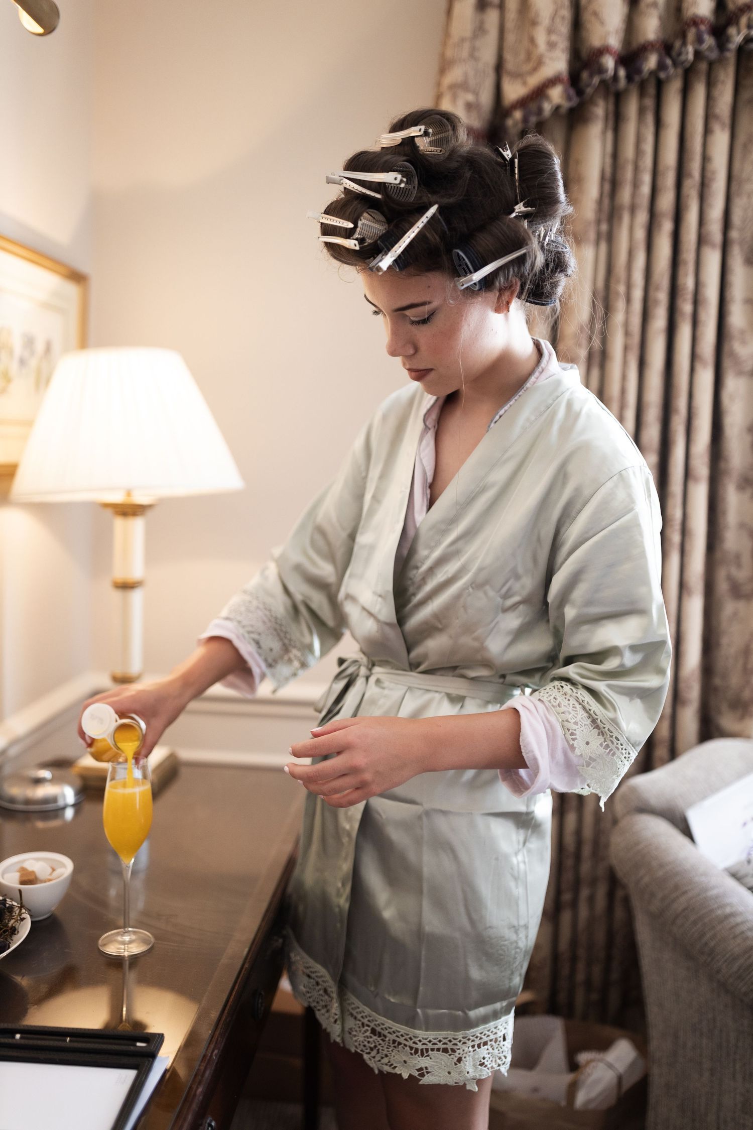 Woman in robe and hair rollers pouring orange juice in an elegant hotel room.