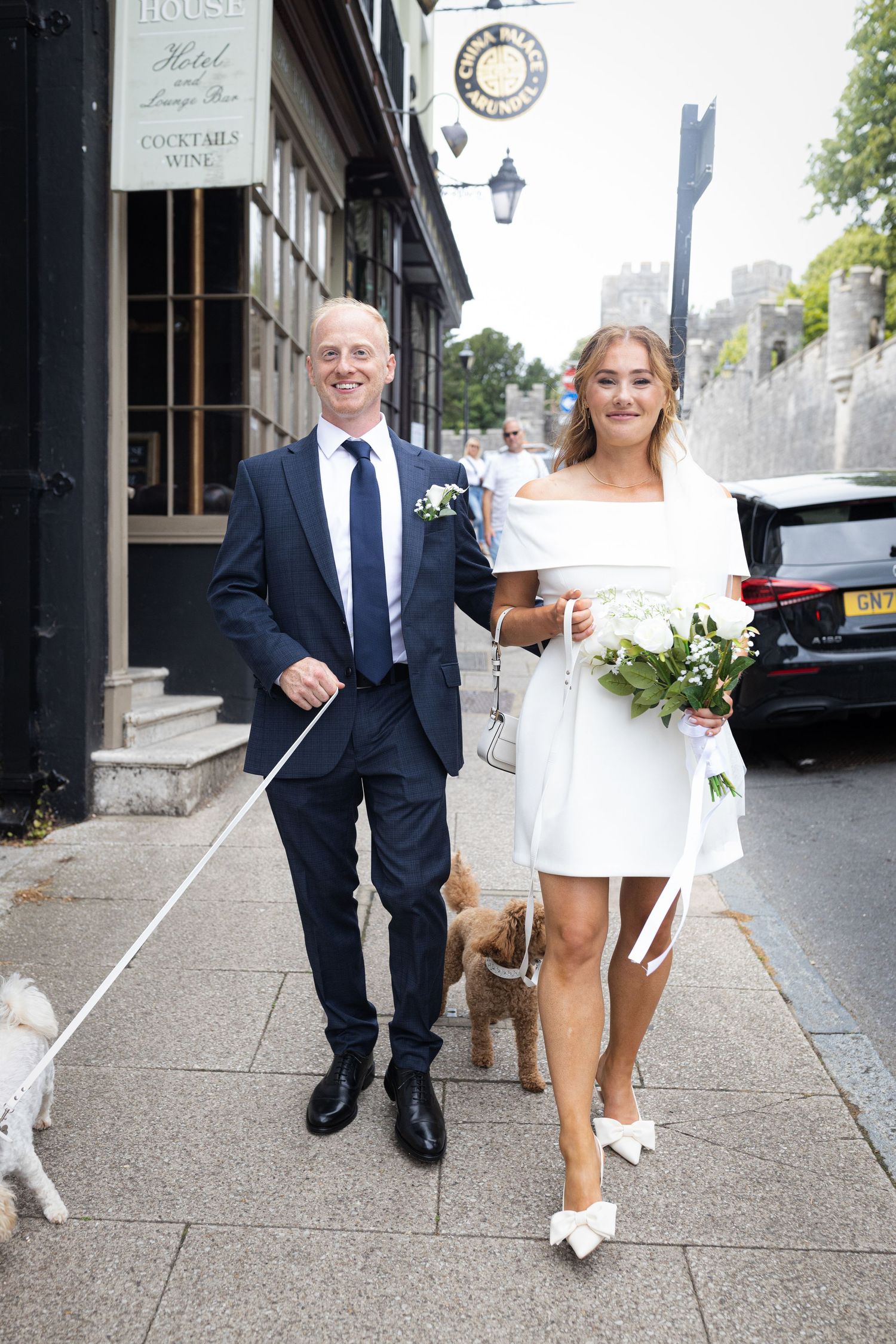 A couple in wedding attire walking their dog on a city sidewalk in front of historic buildings.
