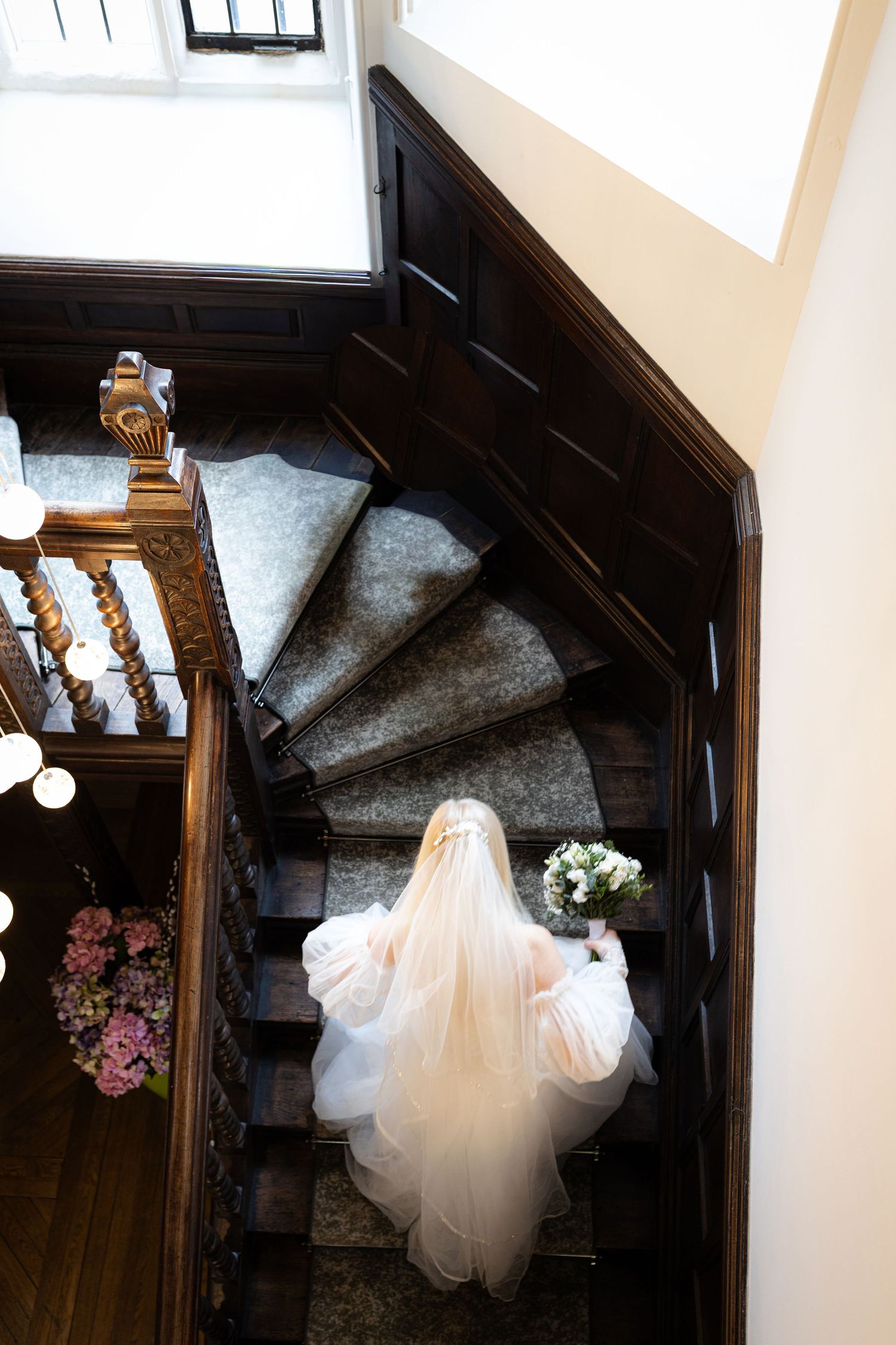 Bride in white gown and veil descends elegant dark wood staircase holding bouquet, viewed from above.