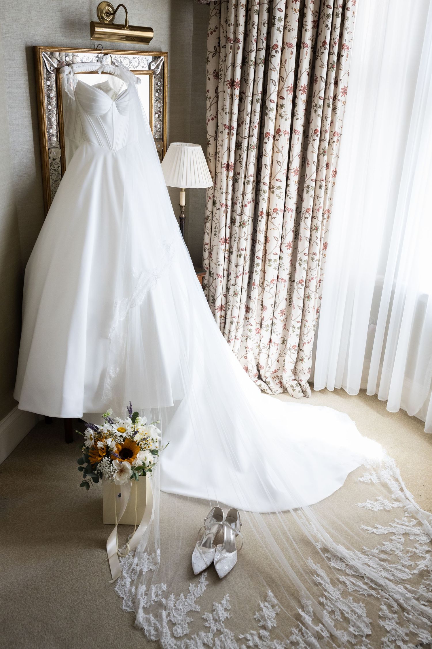 White wedding dress hanging on mirror with lace veil, bridal shoes, and sunflower bouquet in an elegant hotel room.