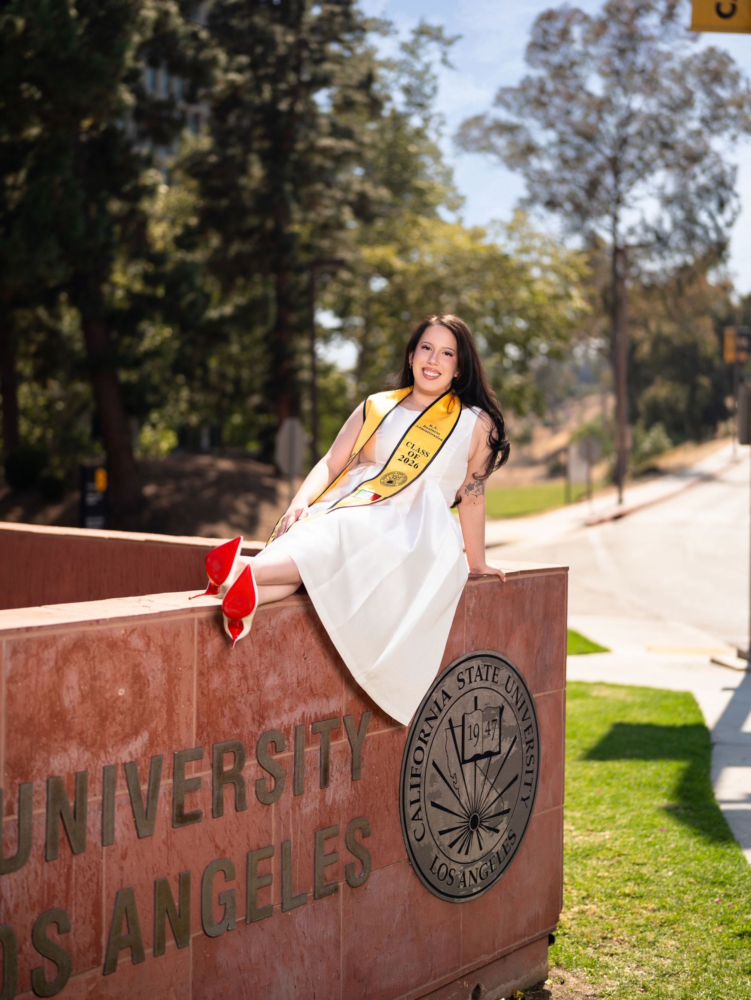 Graduate in white dress and gold stole sits on Cal State LA university sign during graduation photo session.