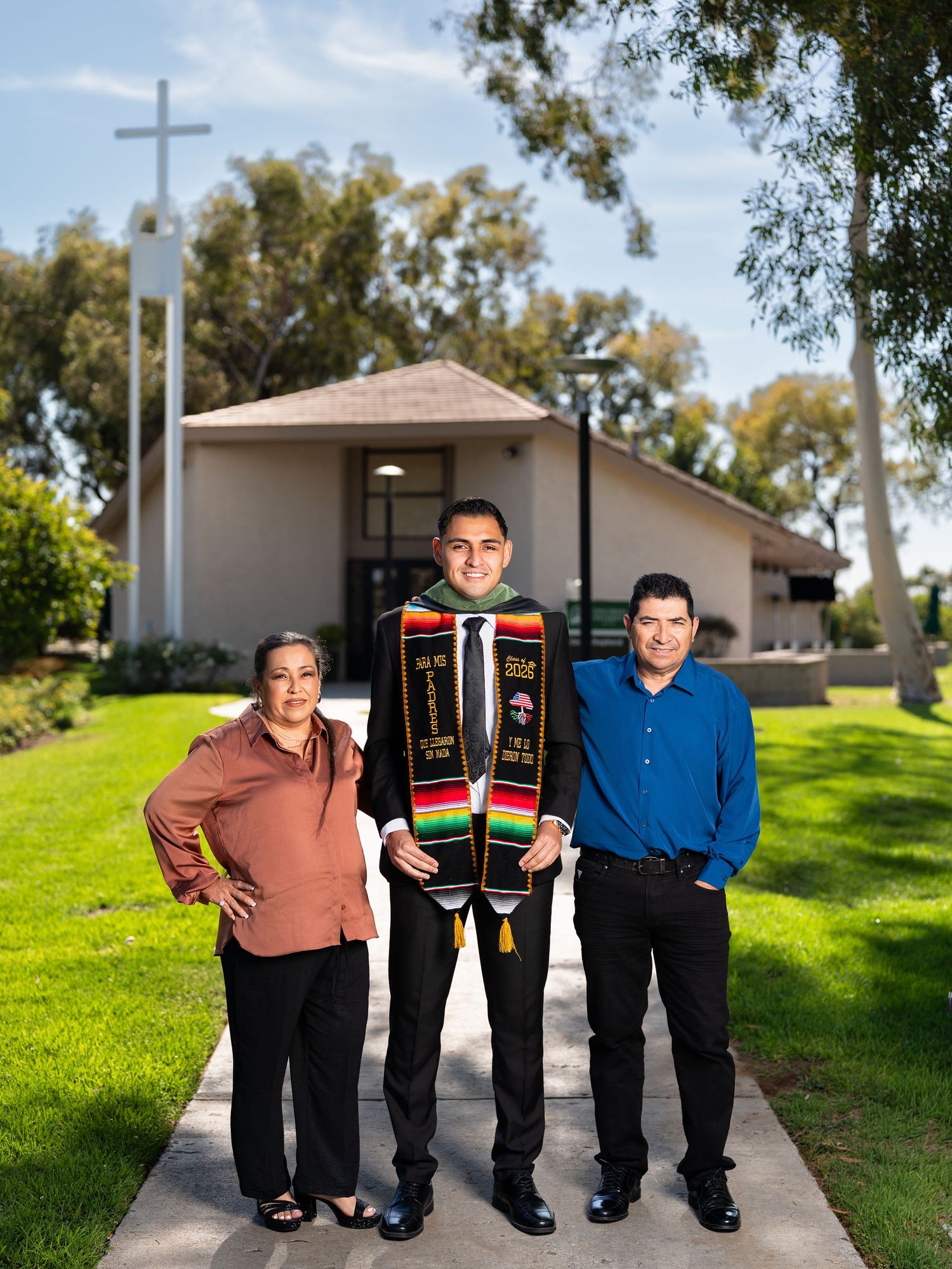 Graduate in cap and gown stole poses with proud family in front of church cross on sunny day.