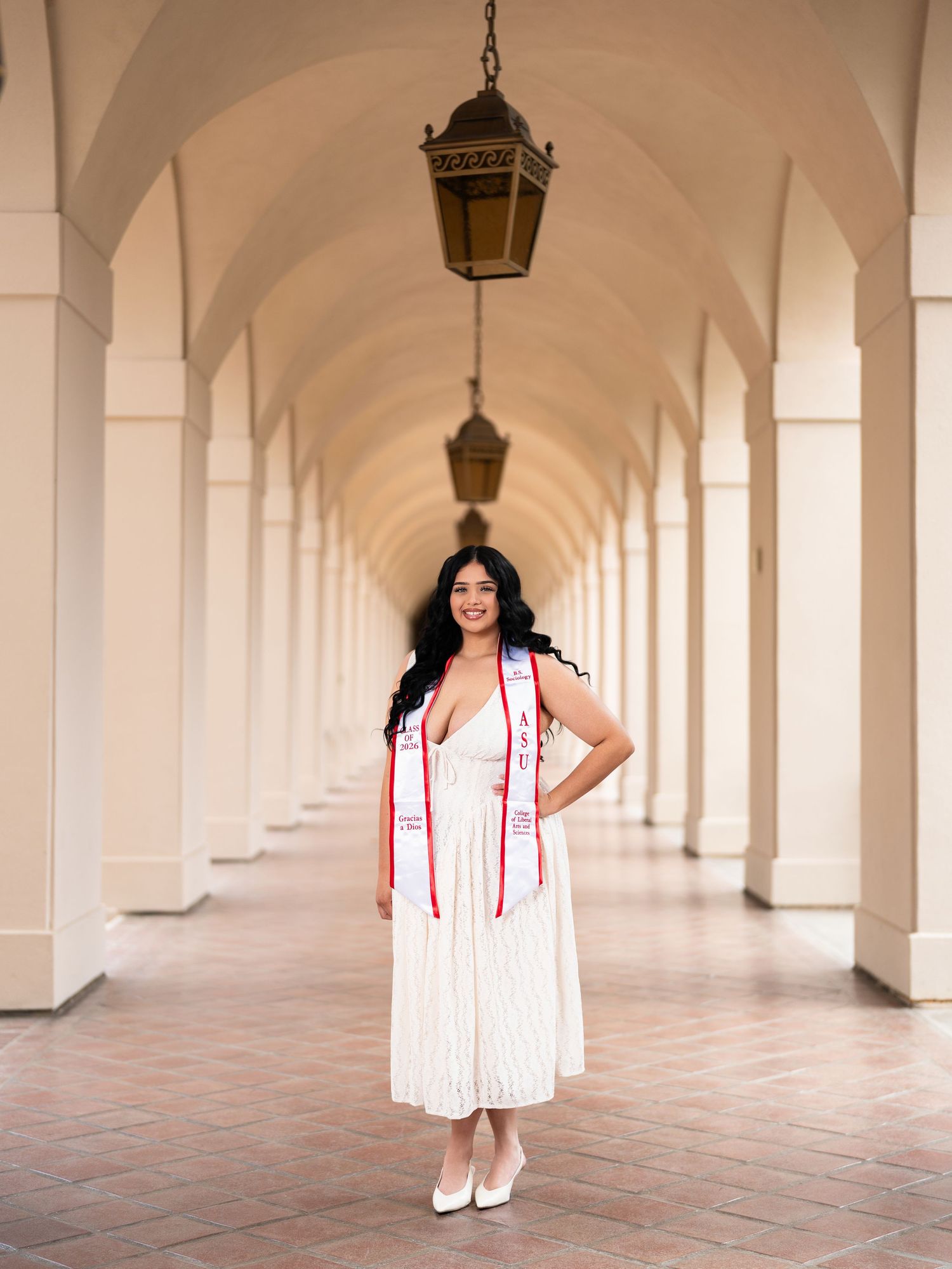 Graduate in white dress and red sash poses under arched colonnade corridor for graduation photos.
