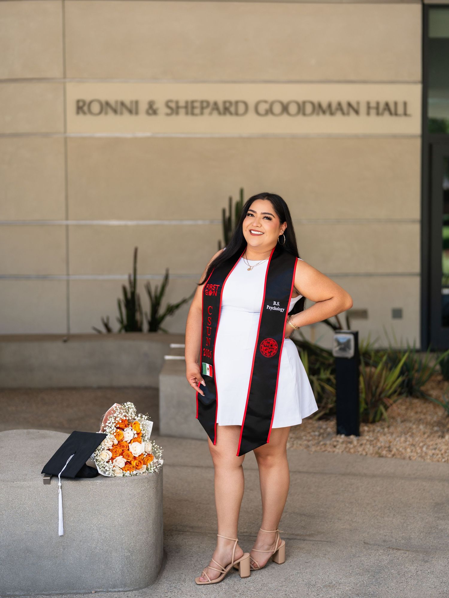 Graduate in white dress and black stole poses proudly before Ronni & Shepard Goodman Hall with flowers and cap.