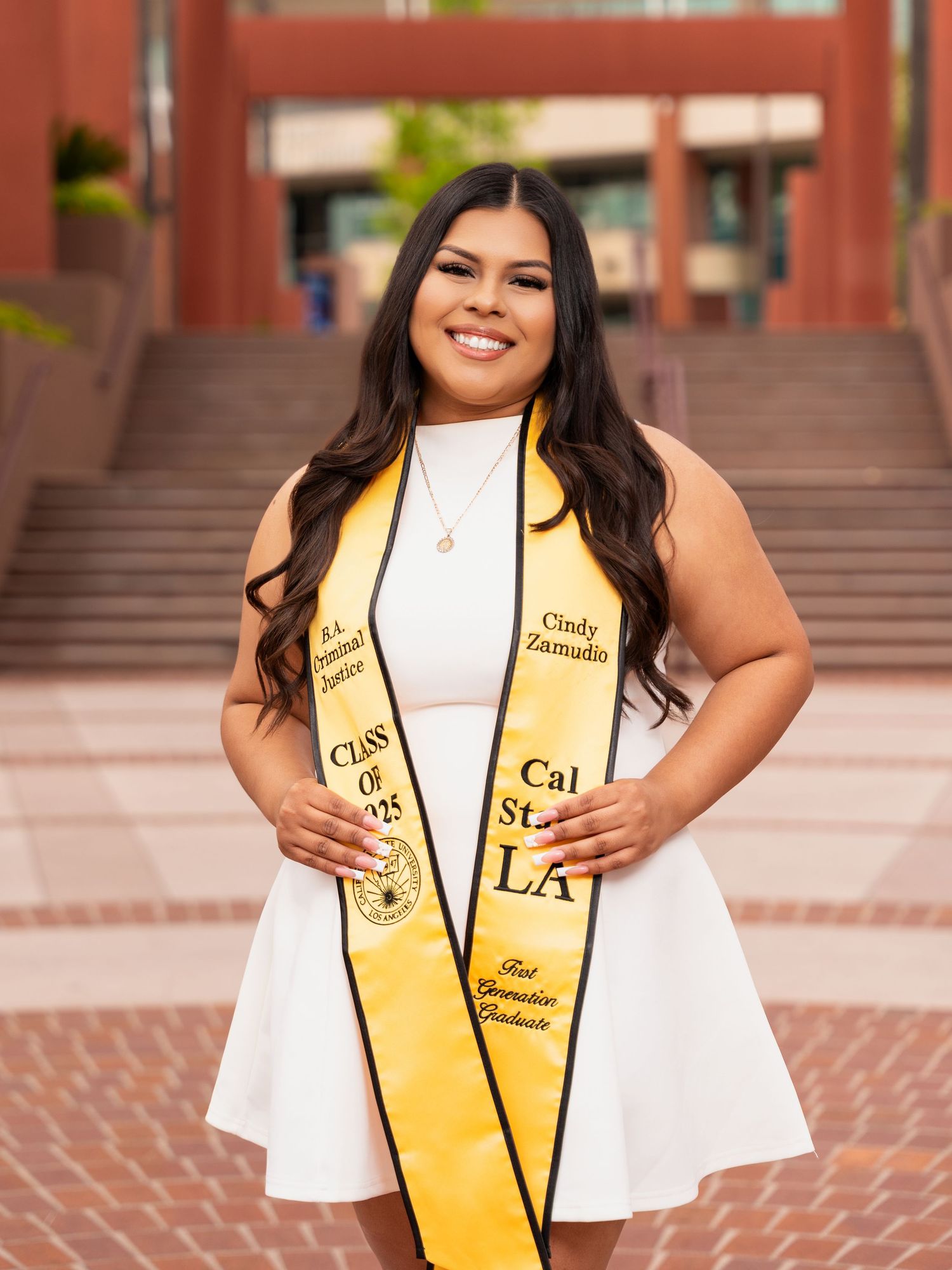 Graduate wearing a white dress and yellow graduation stole with gold trim poses on brick steps outside a building.