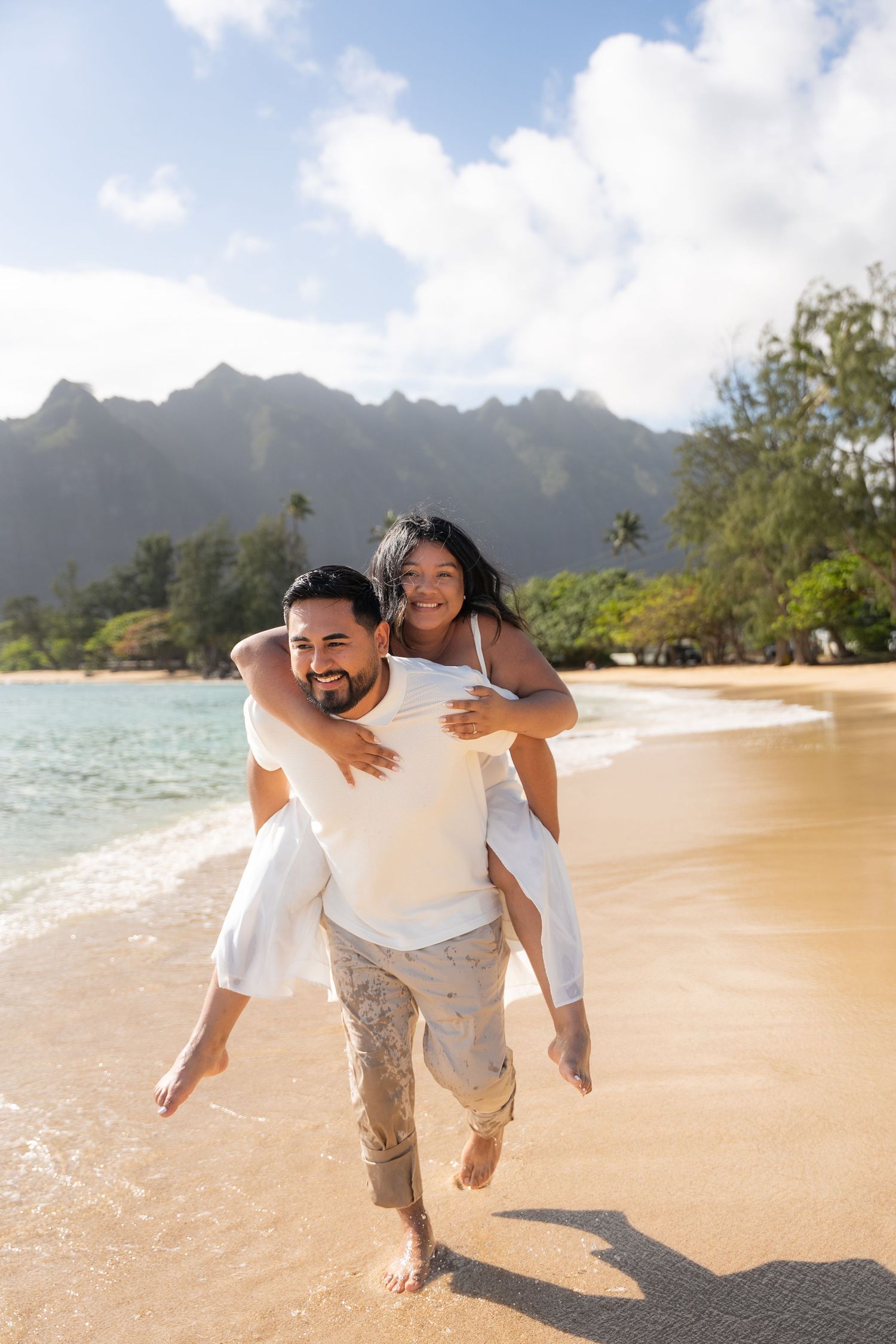 A couple playfully frolics on a scenic Hawaiian beach with blue skies and mountains in the background.