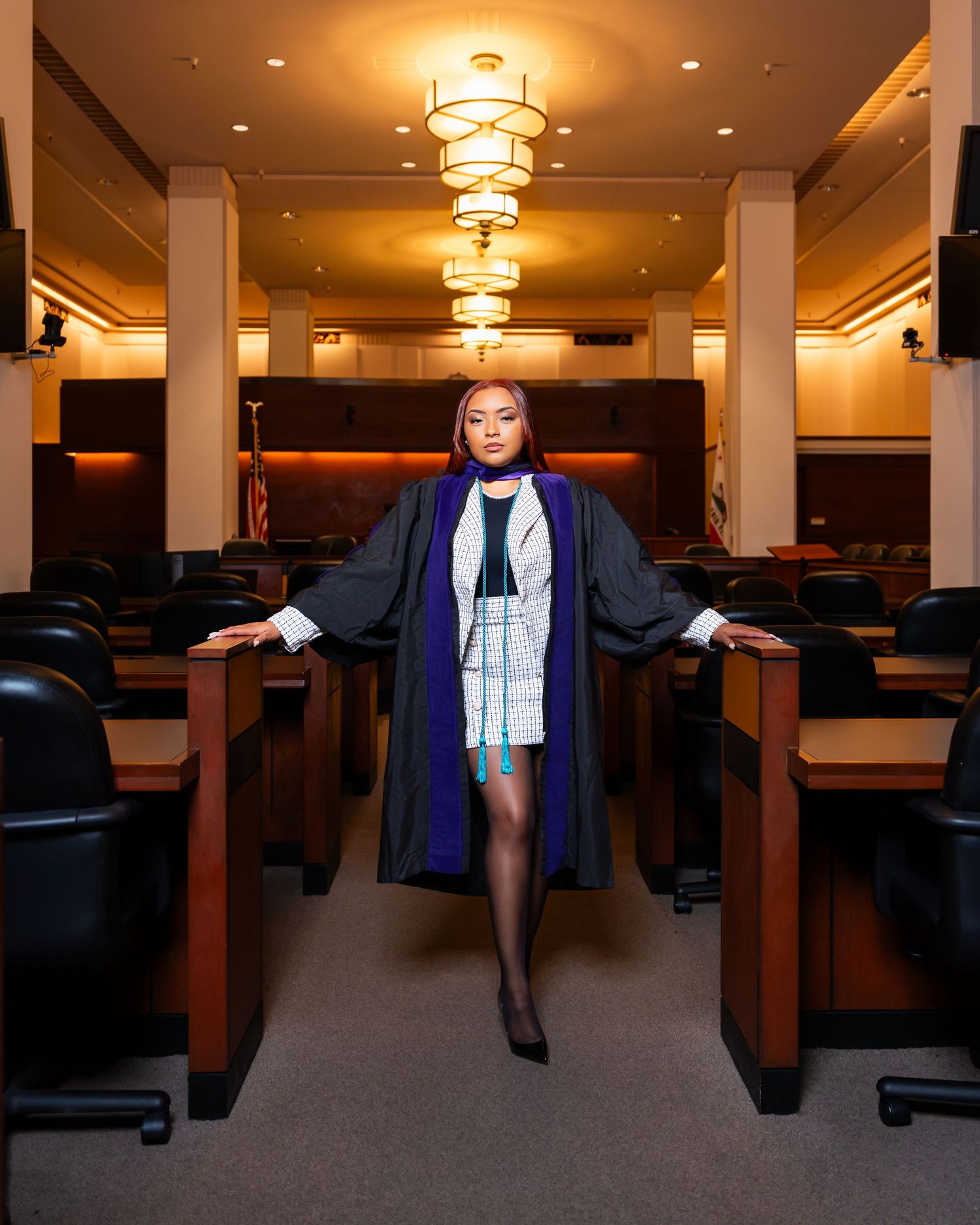 A graduate stands in an auditorium wearing academic regalia and a blue scarf while elegant chandeliers illuminate the space.