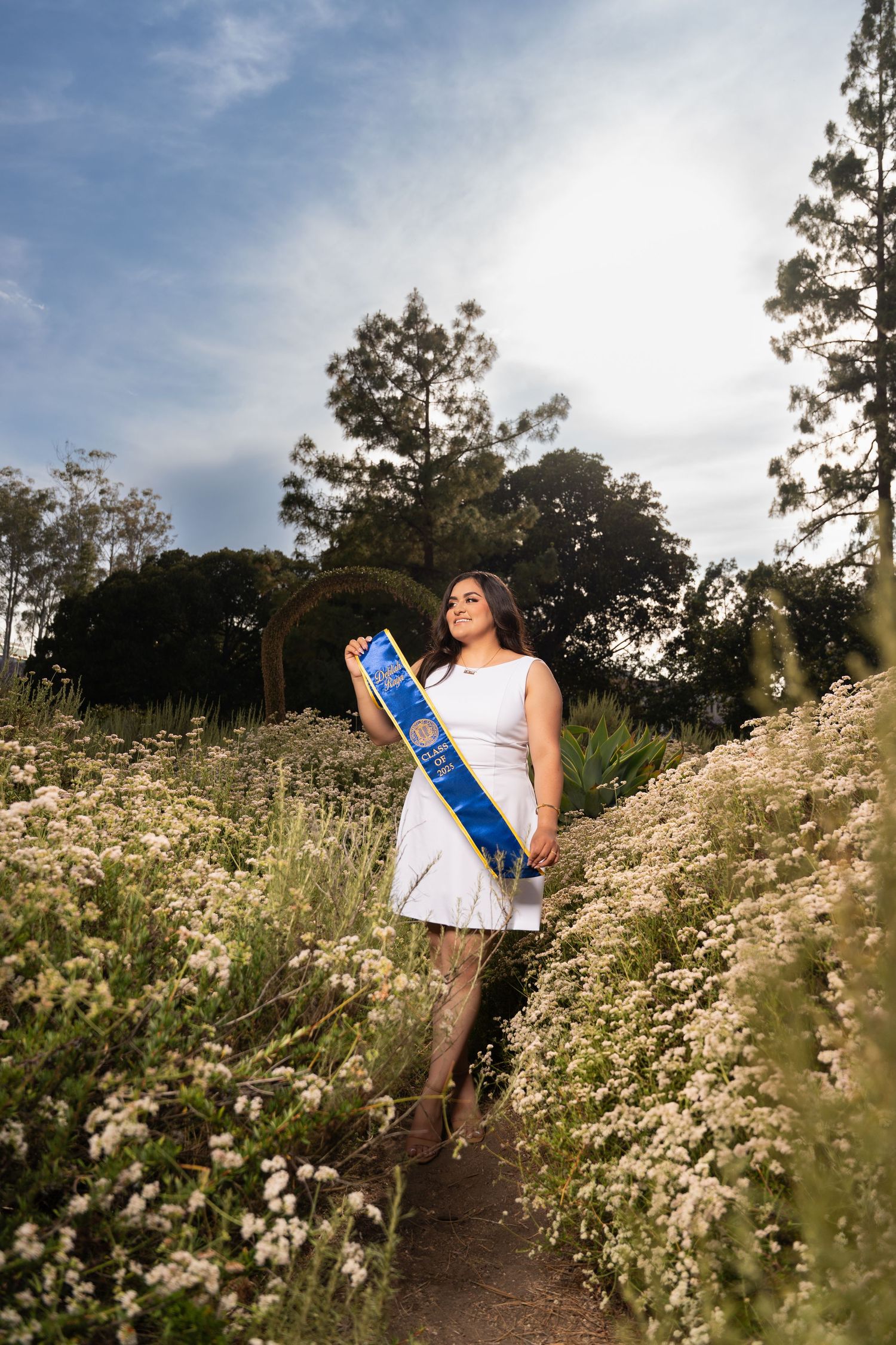 A person wearing a white dress stands on a path surrounded by blooming wildflowers and trees on a sunny day.