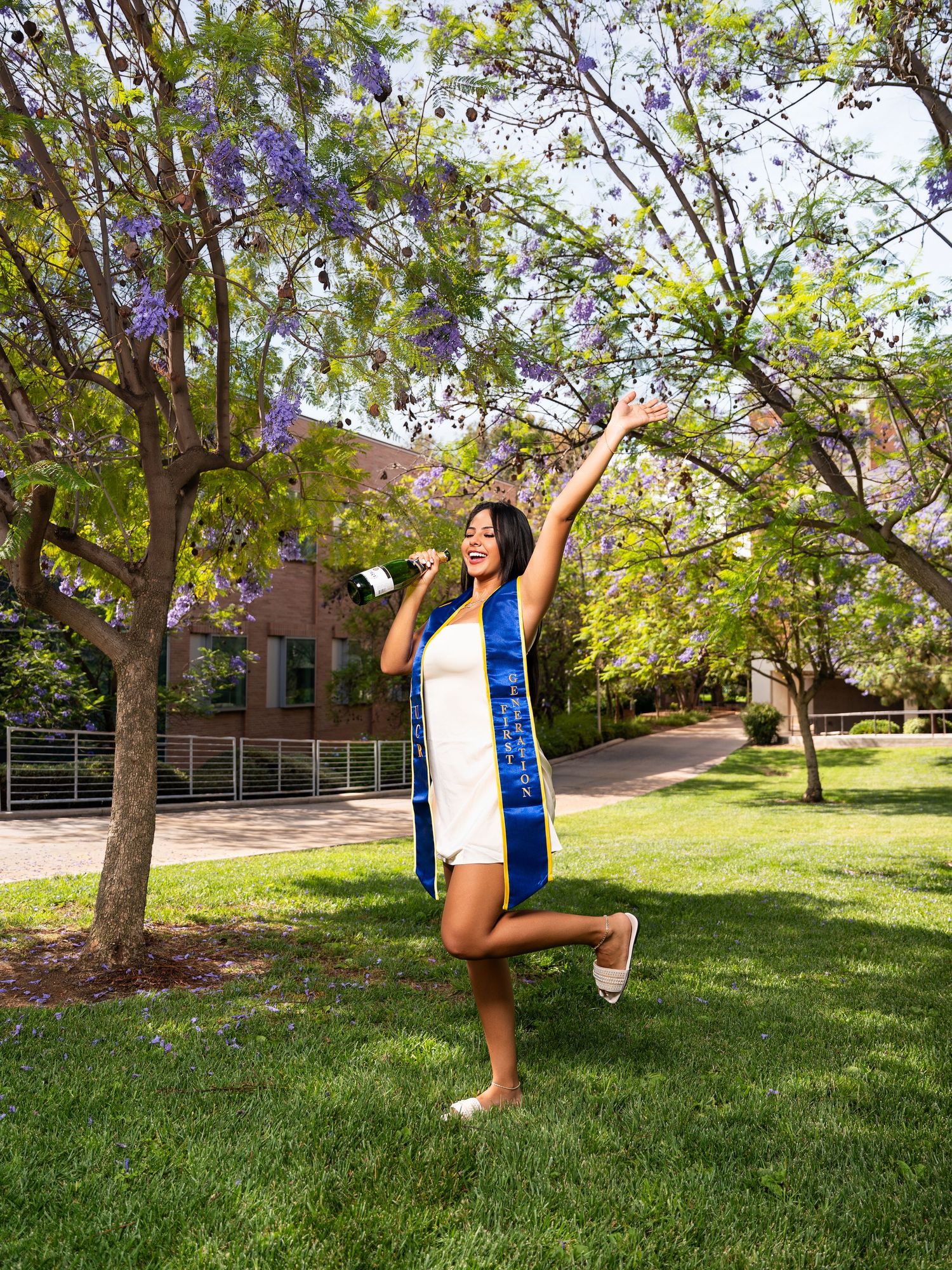 A cheerful graduate in white attire and blue sash poses for celebratory photos on a sunny lawn with trees in the background.