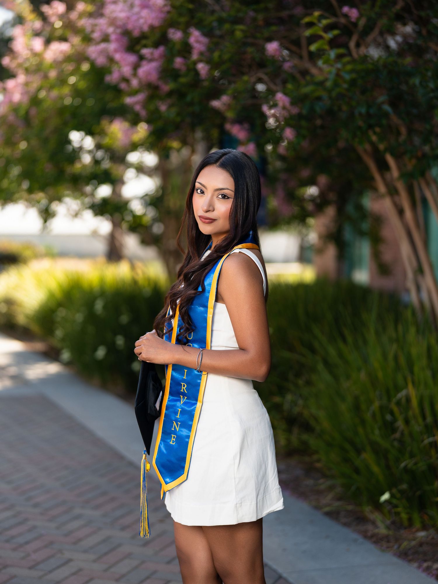 Graduate in white dress and blue gold stole poses outdoors near blooming trees on a sunny day.