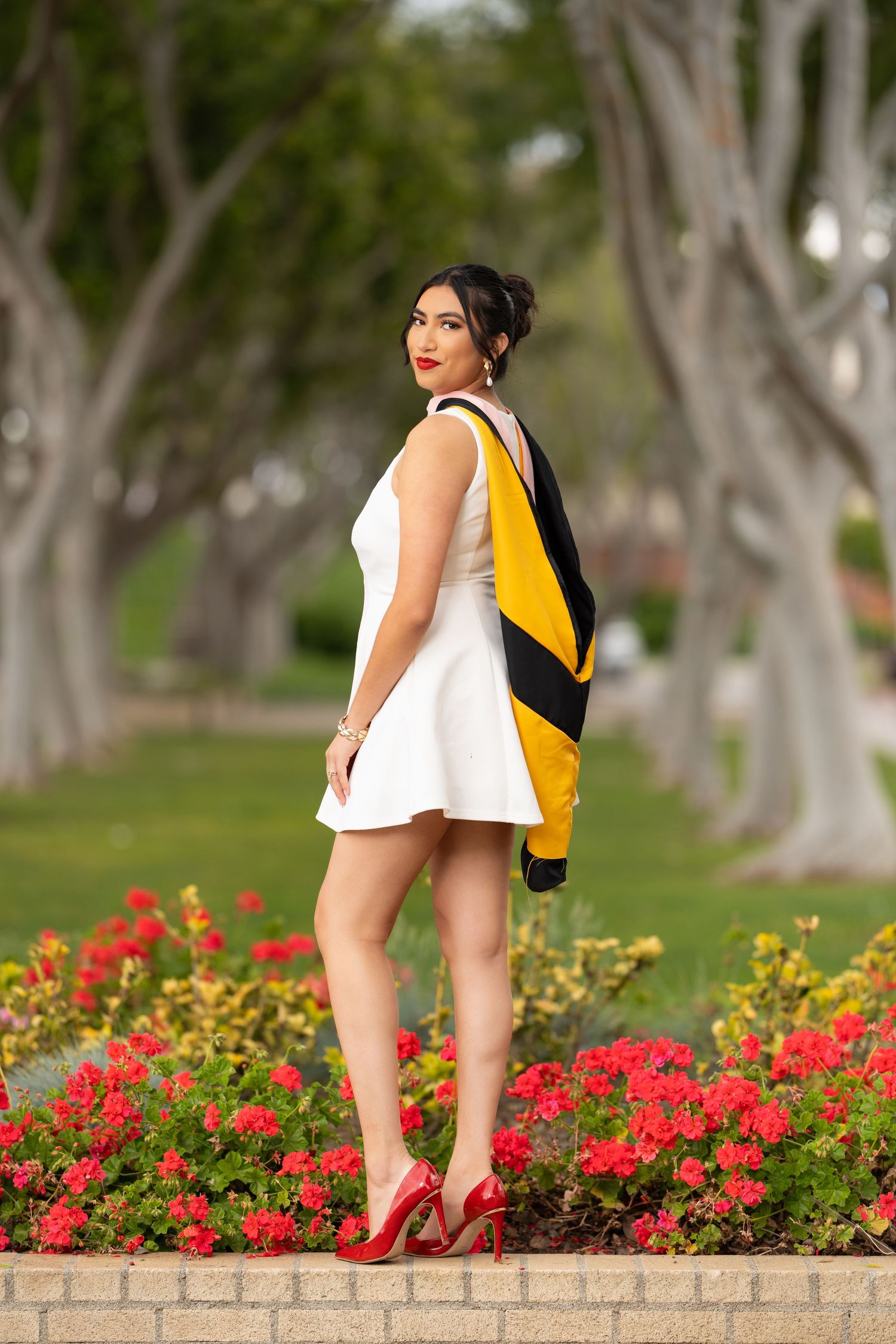 Woman in yellow dress and red heels sits gracefully on stone ledge beside vibrant red and yellow flower garden.